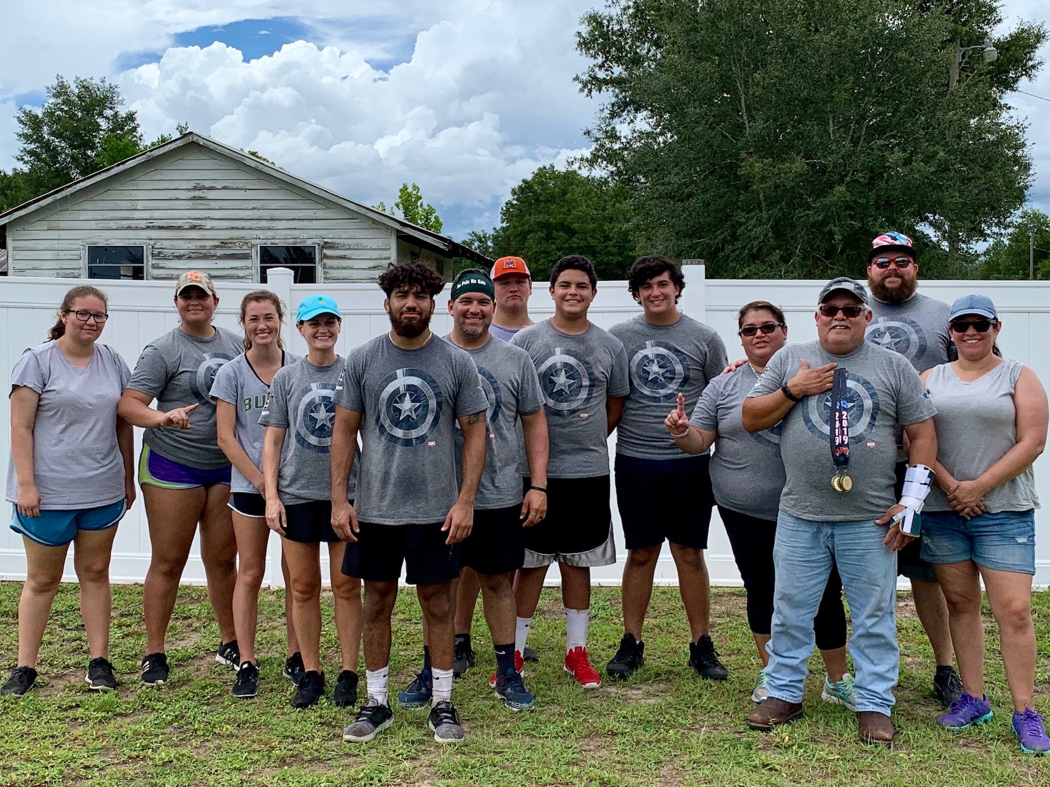 Group of people wearing matching gray shirts with a circular star emblem, posing outdoors in front of a white fence and an old house.