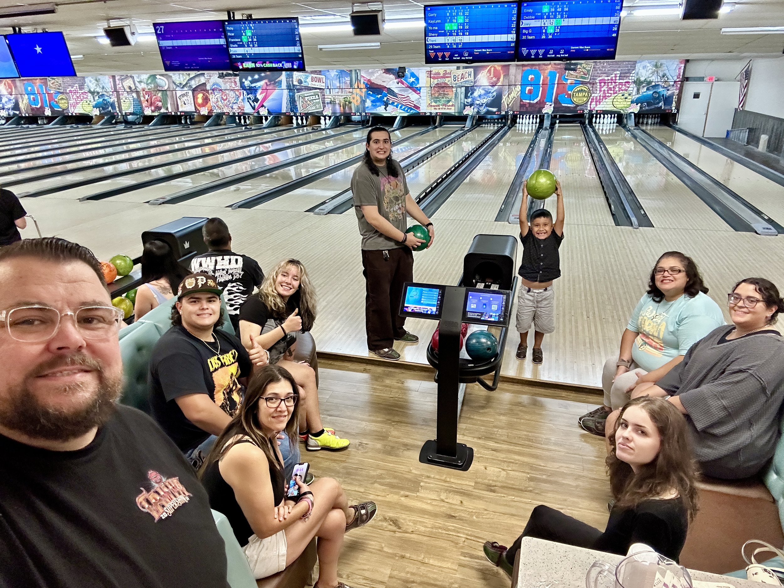 A group of people at a bowling alley, with some sitting on benches and others standing near the bowling lanes. A child is holding a bowling ball and smiling. Bowling balls are visible on the ball return equipment. The background shows multiple bowlin