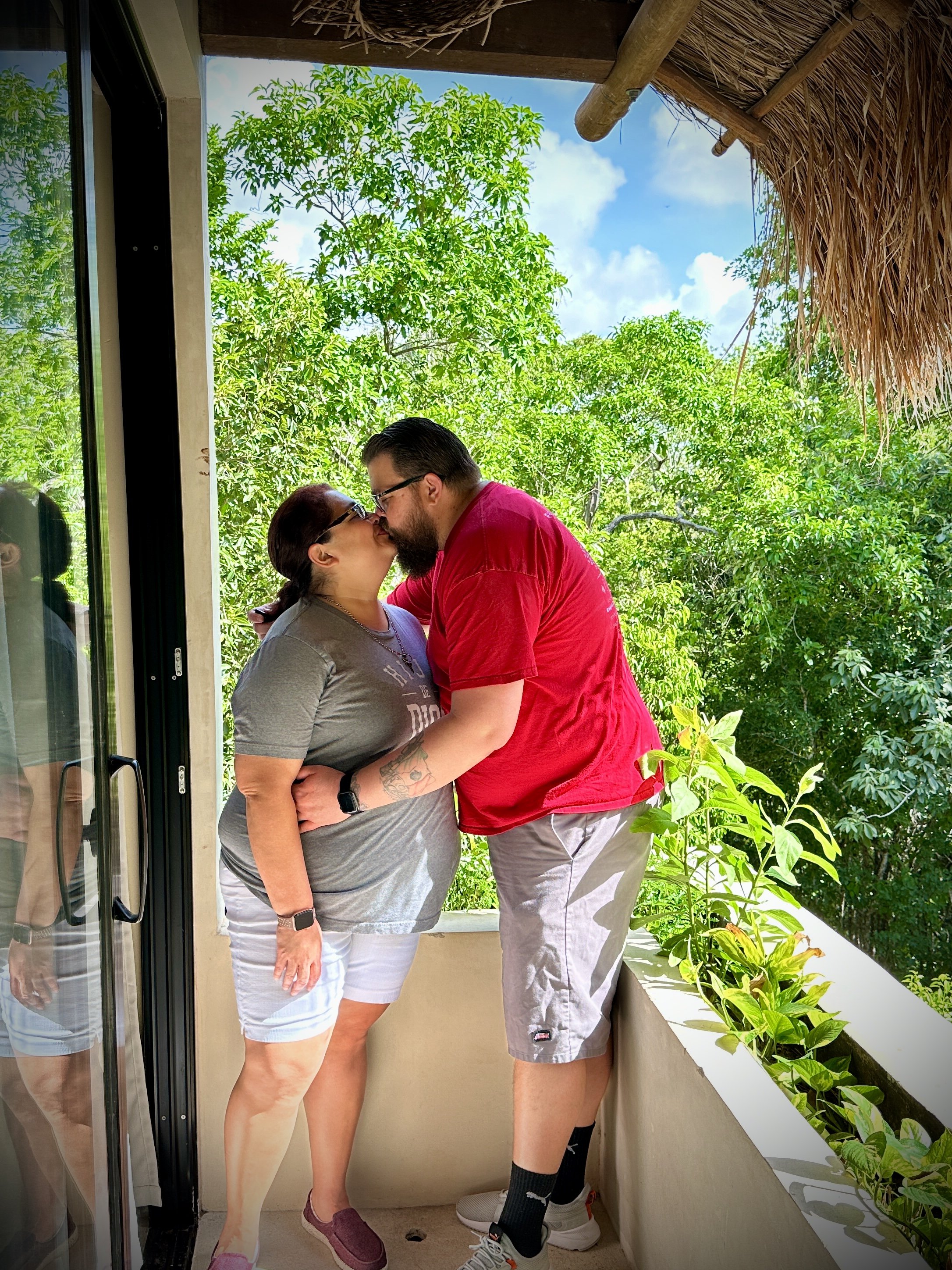 A couple kissing on a balcony with lush green trees in the background. The woman is wearing a gray shirt and white shorts, while the man is wearing a red shirt and gray shorts. The balcony has a thatched roof and a potted plant is visible. Reflection