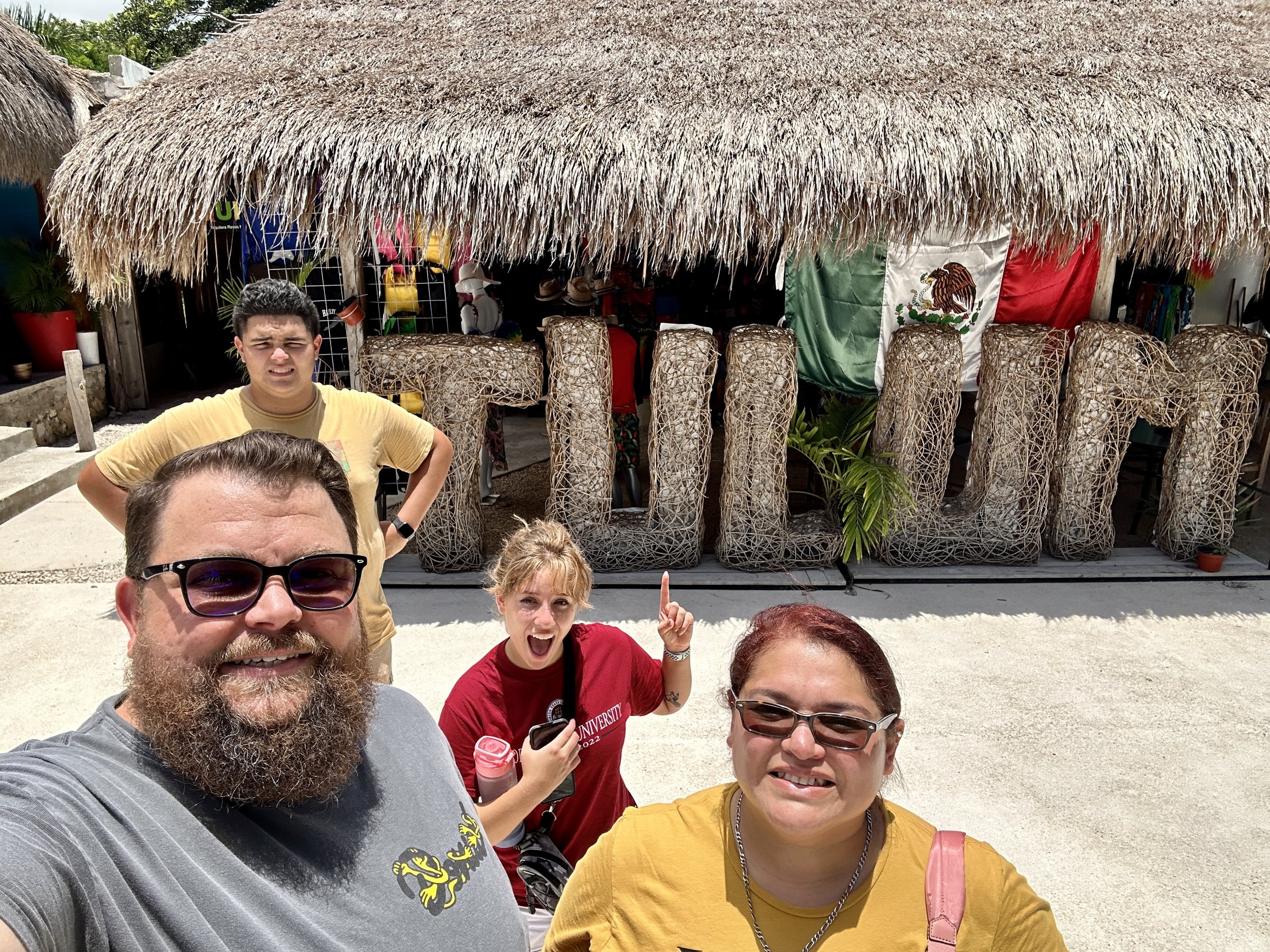 A group of four people standing in front of a thatched-roof shop with a large woven "TULUM" sign and a Mexican flag in the background.