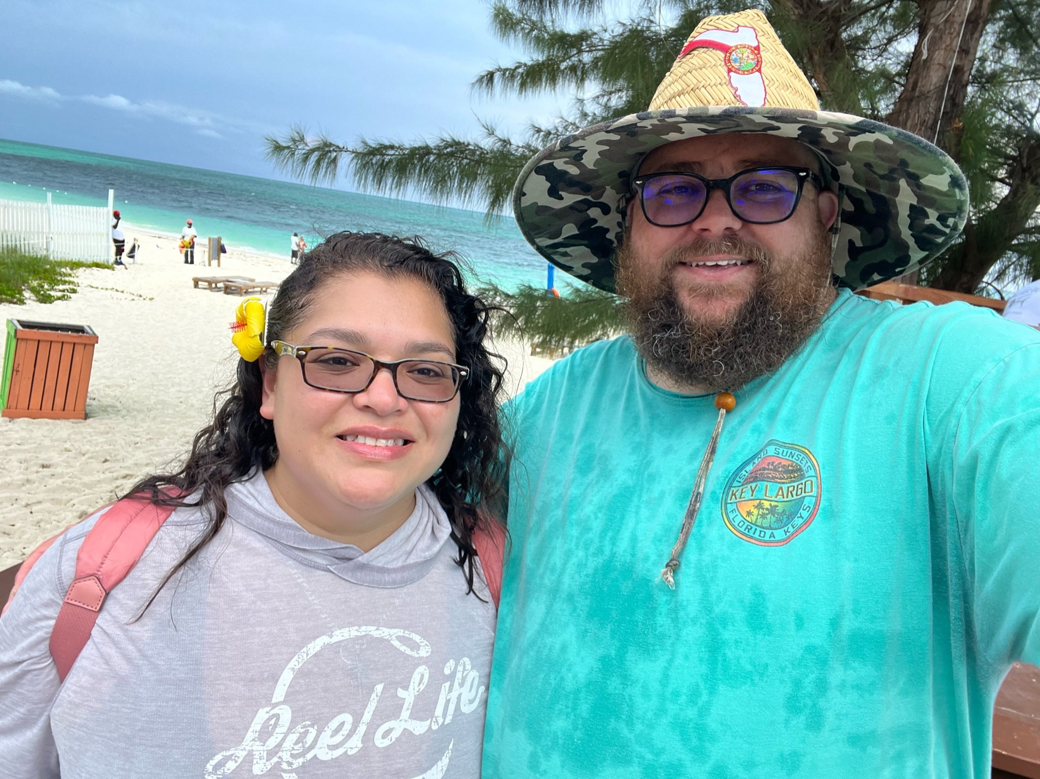 A man and woman smiling at the beach with the ocean and trees in the background, the man wearing a straw hat and glasses, and the woman with a flower in her hair.