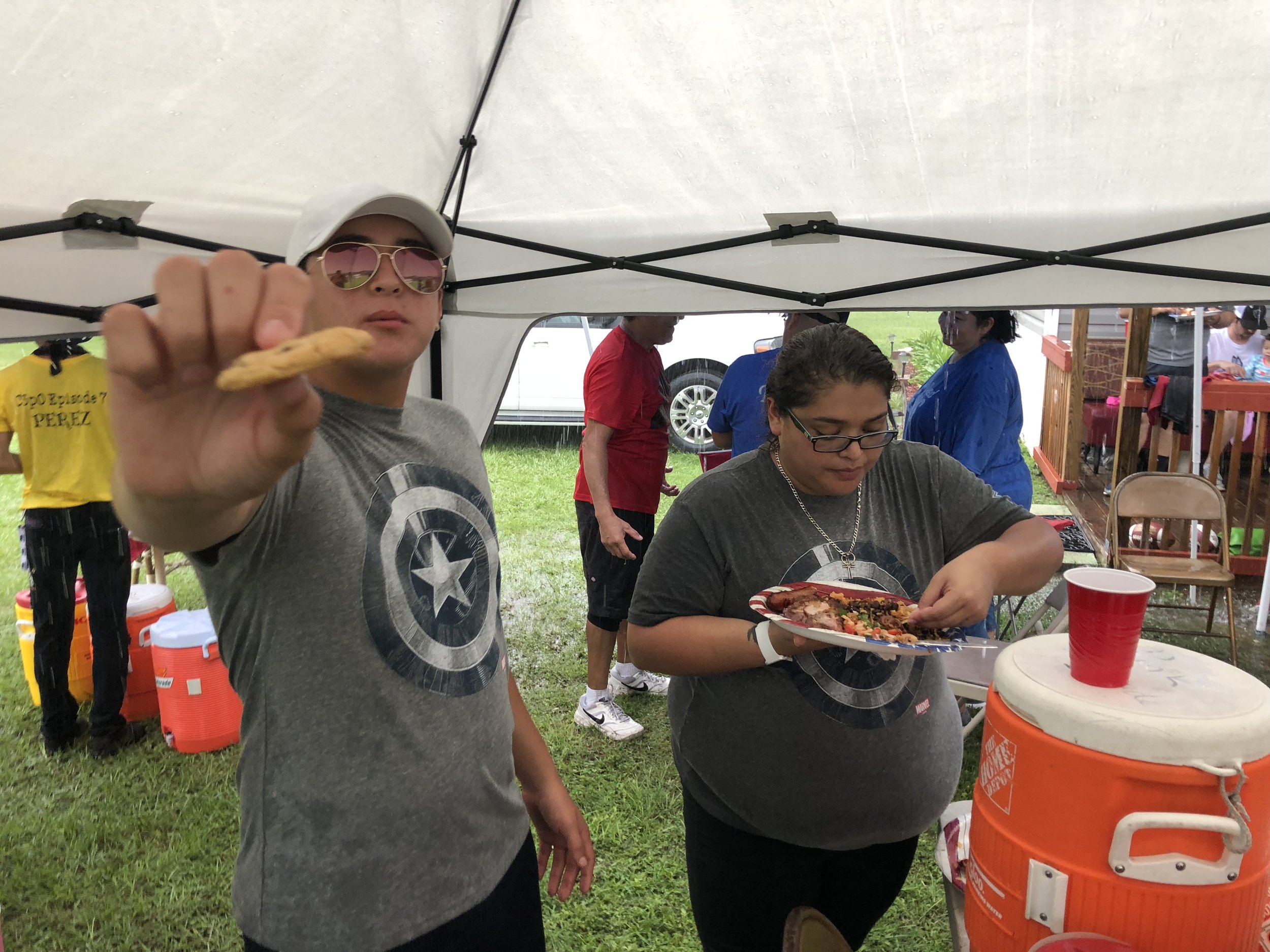 People under a canopy at an outdoor event; one person holding a cookie, another handling a plate of food near an orange cooler.
