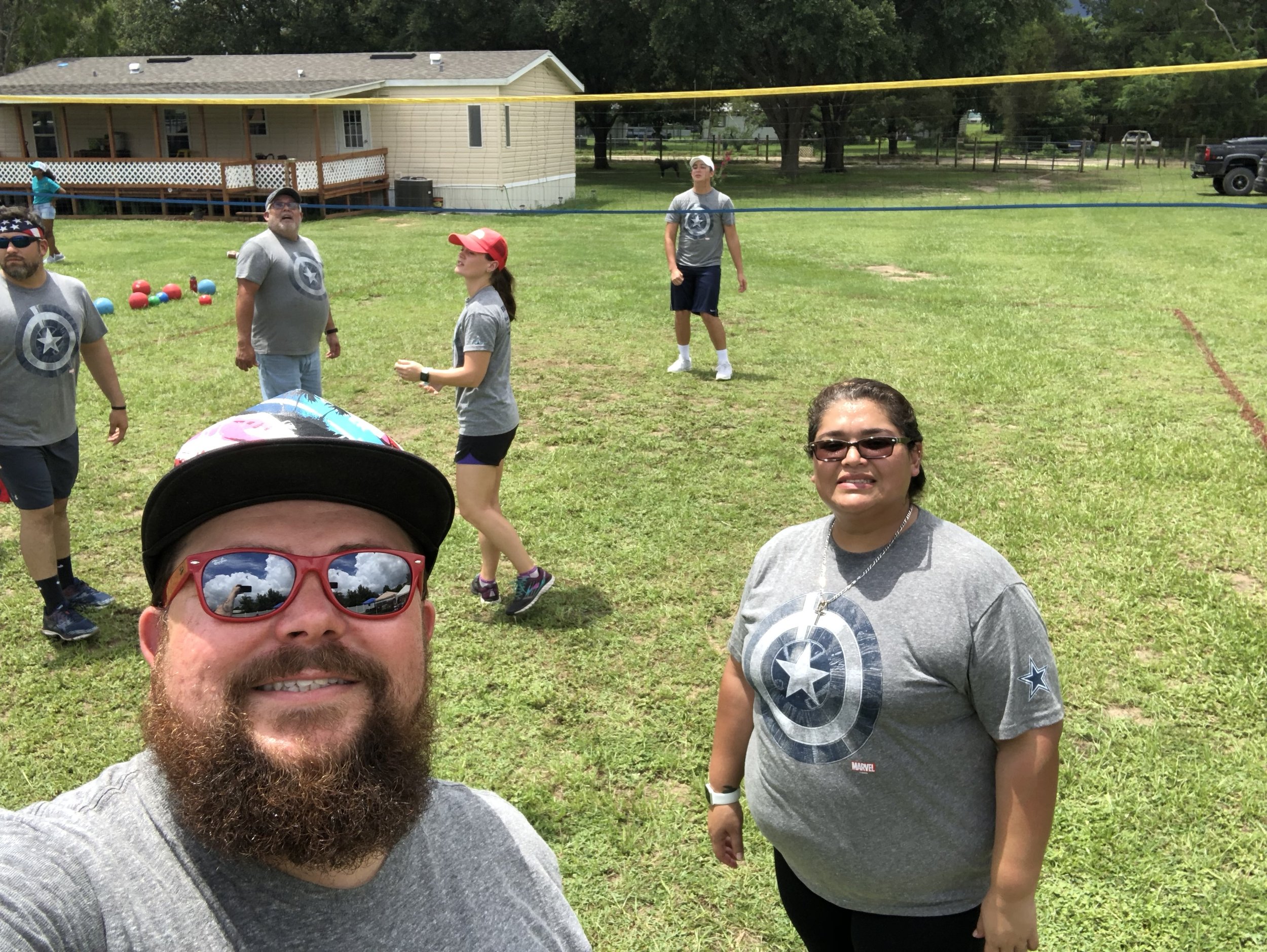 A group of people playing volleyball outdoors on a grassy field, wearing matching gray T-shirts with a circular emblem. One person in the foreground is taking a selfie, wearing sunglasses and a hat. There's a single-story house and trees in the backg