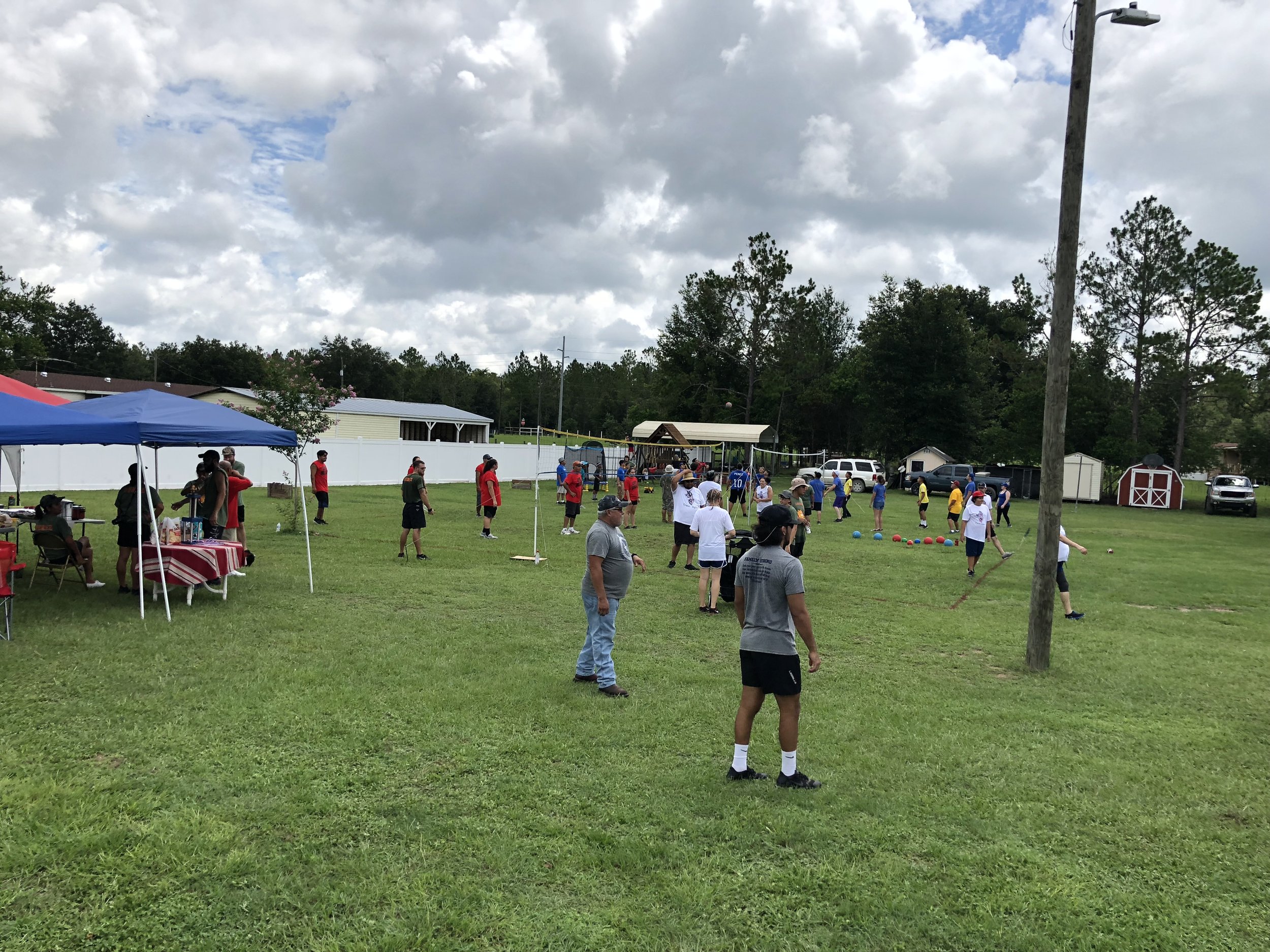 Outdoor event with people playing and socializing on a grassy field. There are tents, sports activities, and casual gatherings. Trees and parked cars are in the background.
