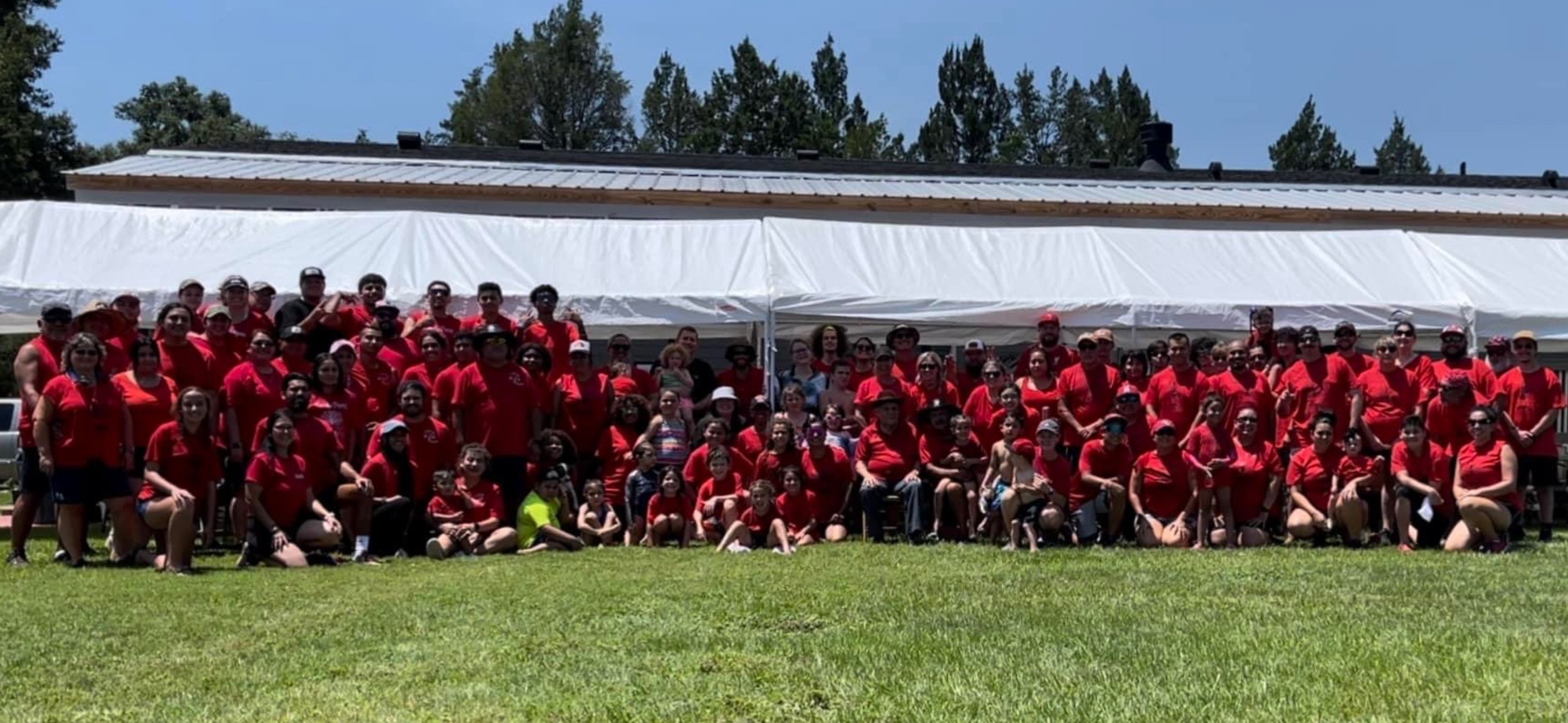 Large group of people wearing red shirts posing outdoors in front of a white tent and building.