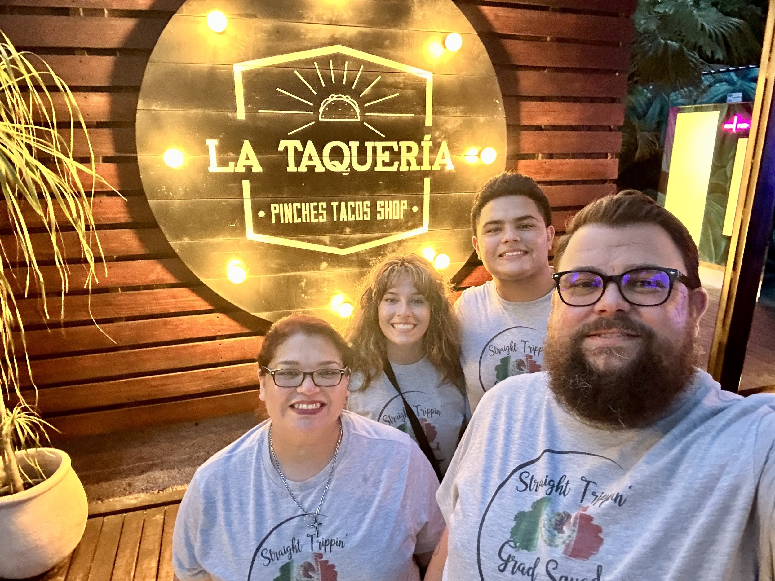 Four people wearing matching "Straight Trippin' Grad Squad" shirts pose in front of a lighted sign for "La Taquería Pinches Tacos Shop" on a wooden wall.
