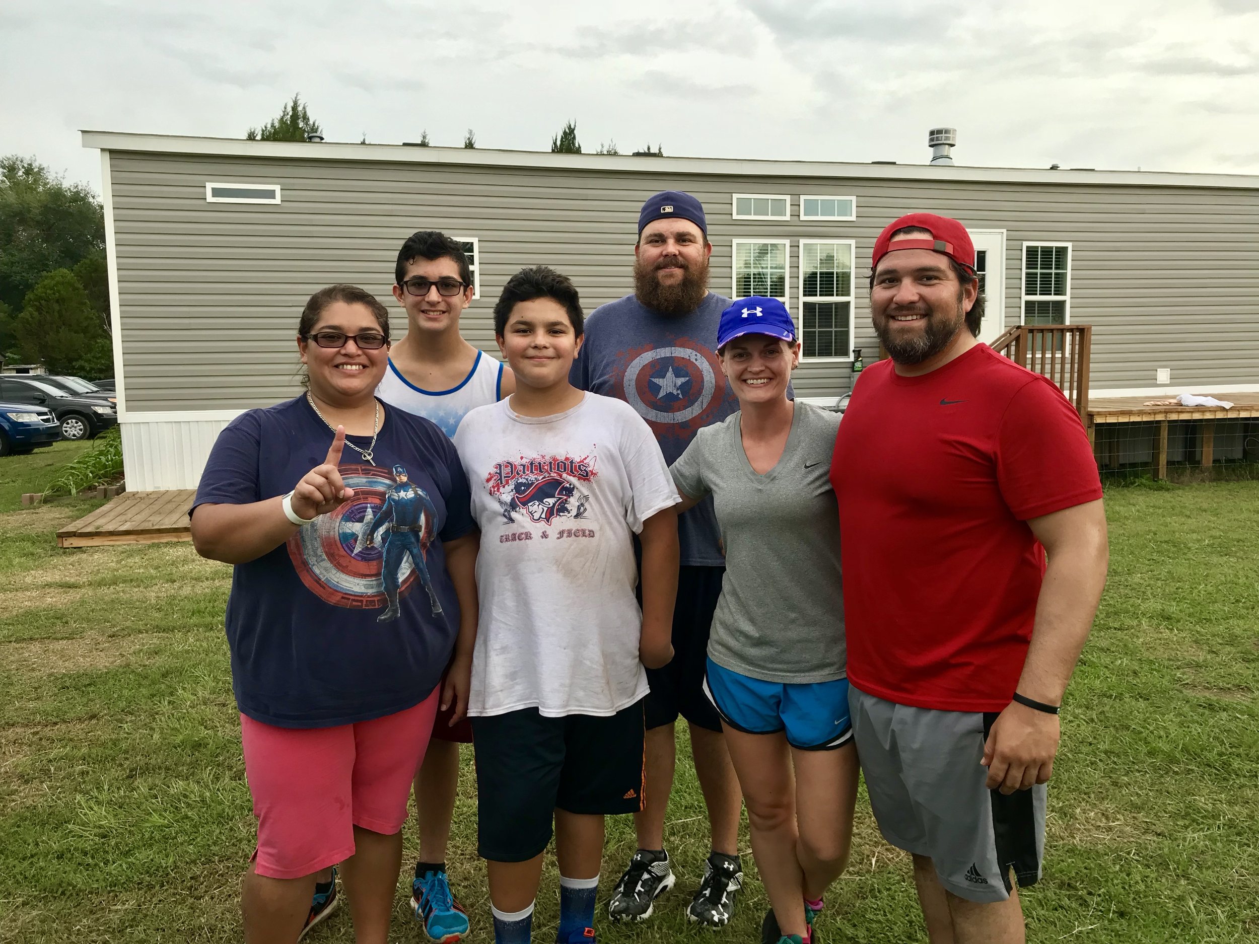 Group of six people smiling in athletic wear outside a house, grass field, overcast sky.