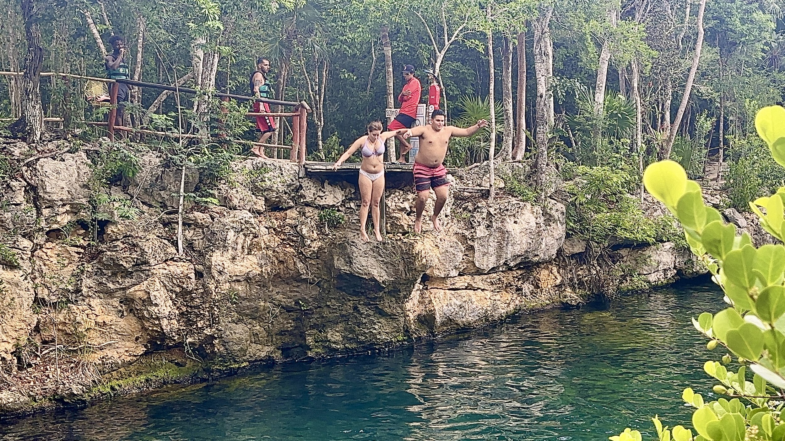 Two people jumping into water from a rocky ledge surrounded by trees, with onlookers watching from above.