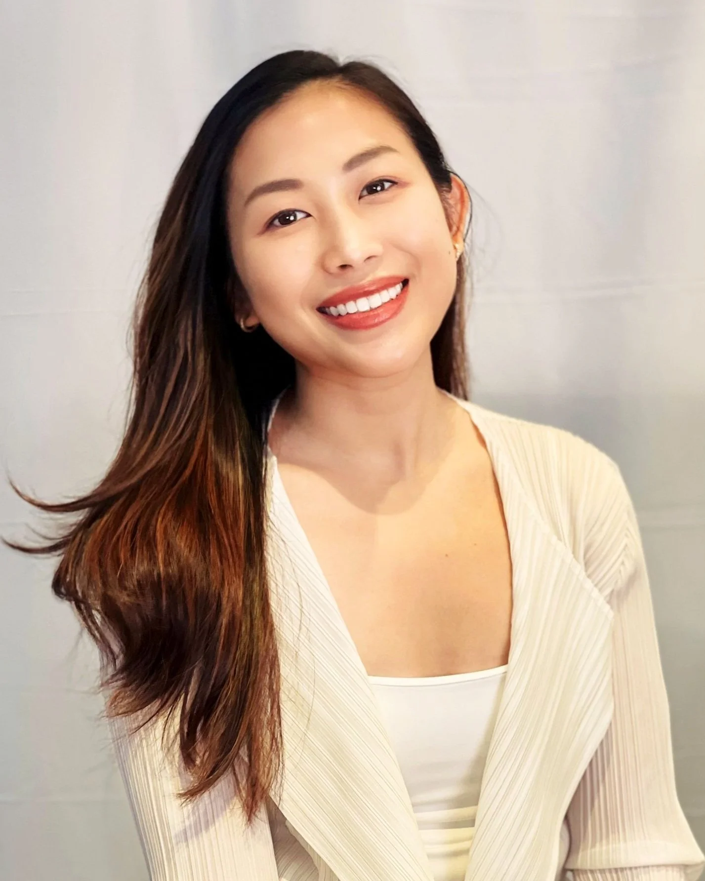 A smiling woman with long dark hair wearing a cream-colored blazer and white top, standing against a plain background.