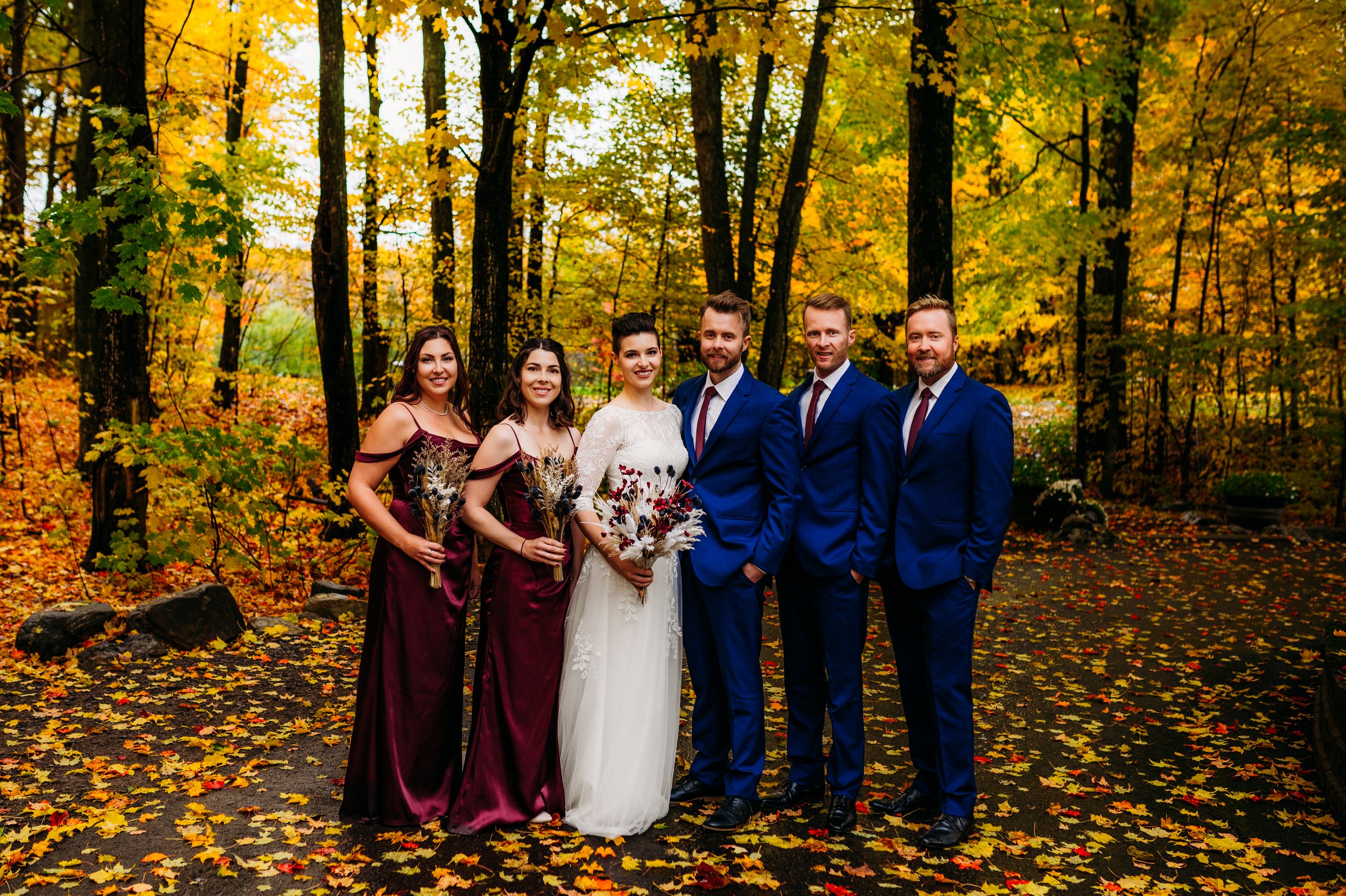 Wedding party posing in fall forest with bride, bridesmaids in burgundy dresses, and groomsmen in blue suits.