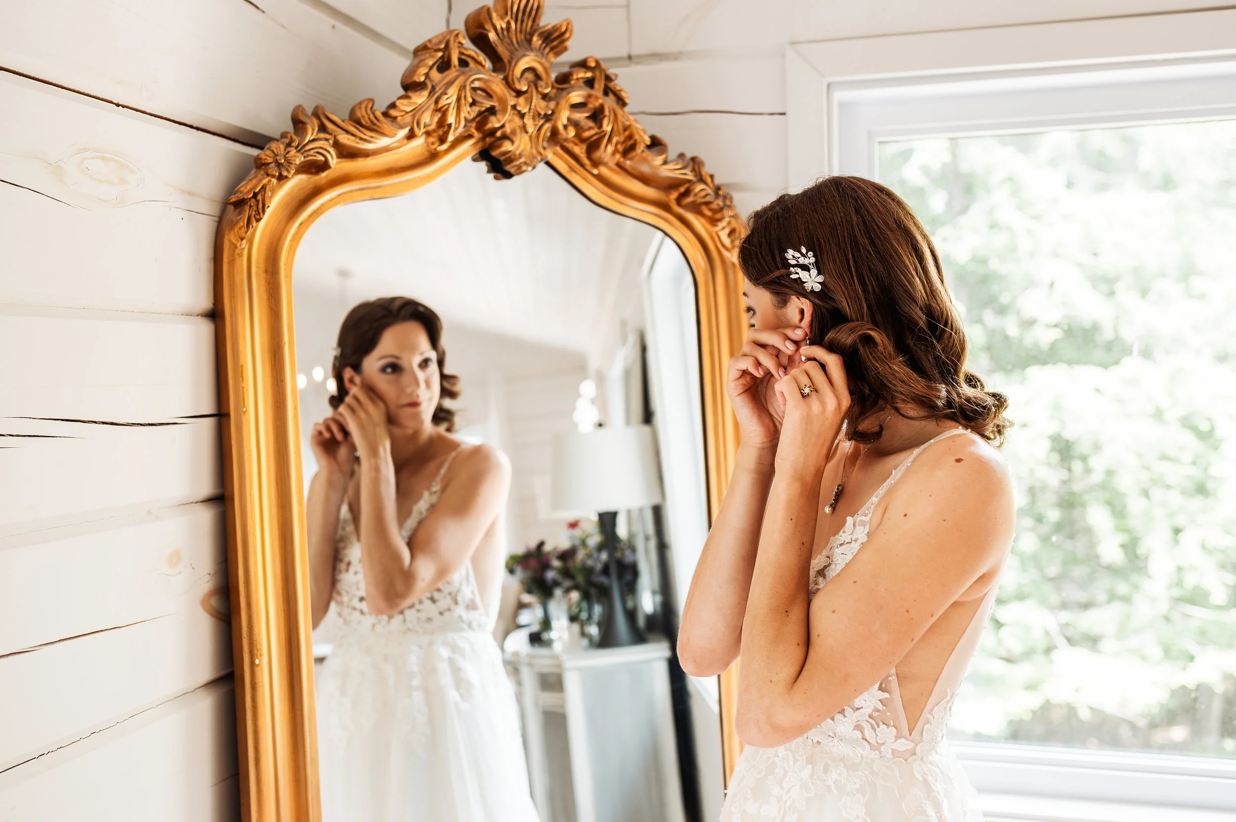 Bride putting on earrings in front of a gold-framed mirror at Le Belvédère, with her reflection showing her lace wedding gown and delicate bridal hairpiece.