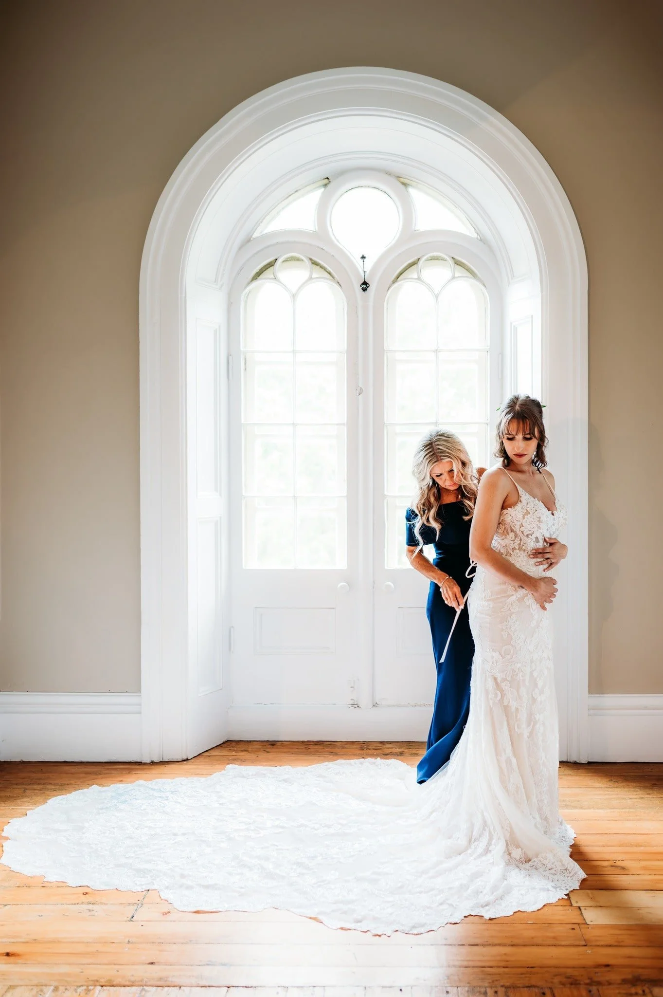 A bride stands by a tall window at Stratmere while the mother of the bride helps tie her wedding dress on the morning of the wedding.