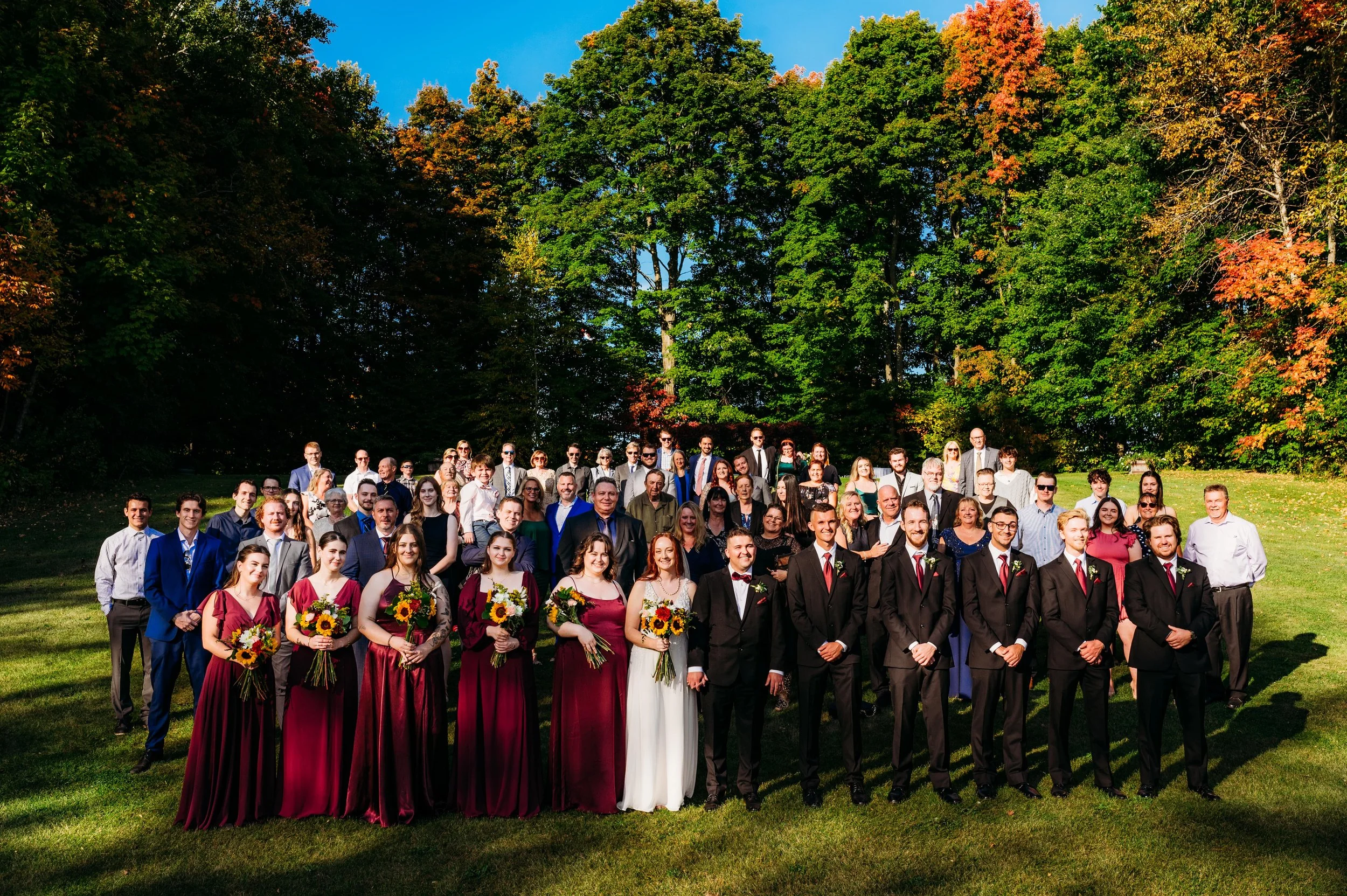Formal wedding group posing outdoors with trees in background, bridesmaids in burgundy dresses holding sunflowers, groomsmen in black suits, and guests standing behind.
