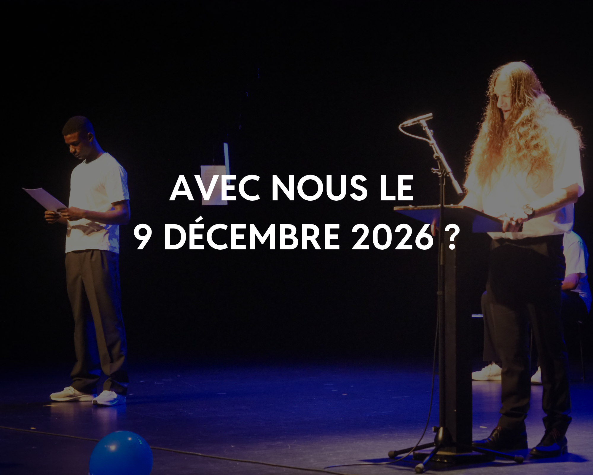 Deux personnes sur scène, une homme et une femme, en train de lire ou de parler lors d'un événement. La scène est sombre avec un éclairage concentré sur eux. Un microphone est visible en face de la femme. Il y a des ballons bleus sur le sol.