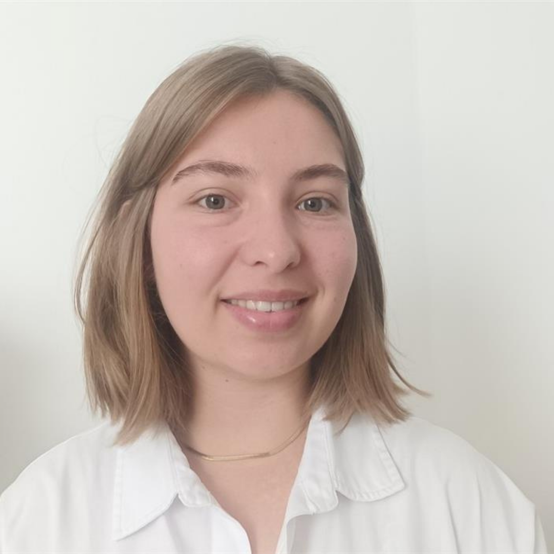 Une jeune femme souriante avec des cheveux mi-longs et une chemise blanche devant un mur blanc.