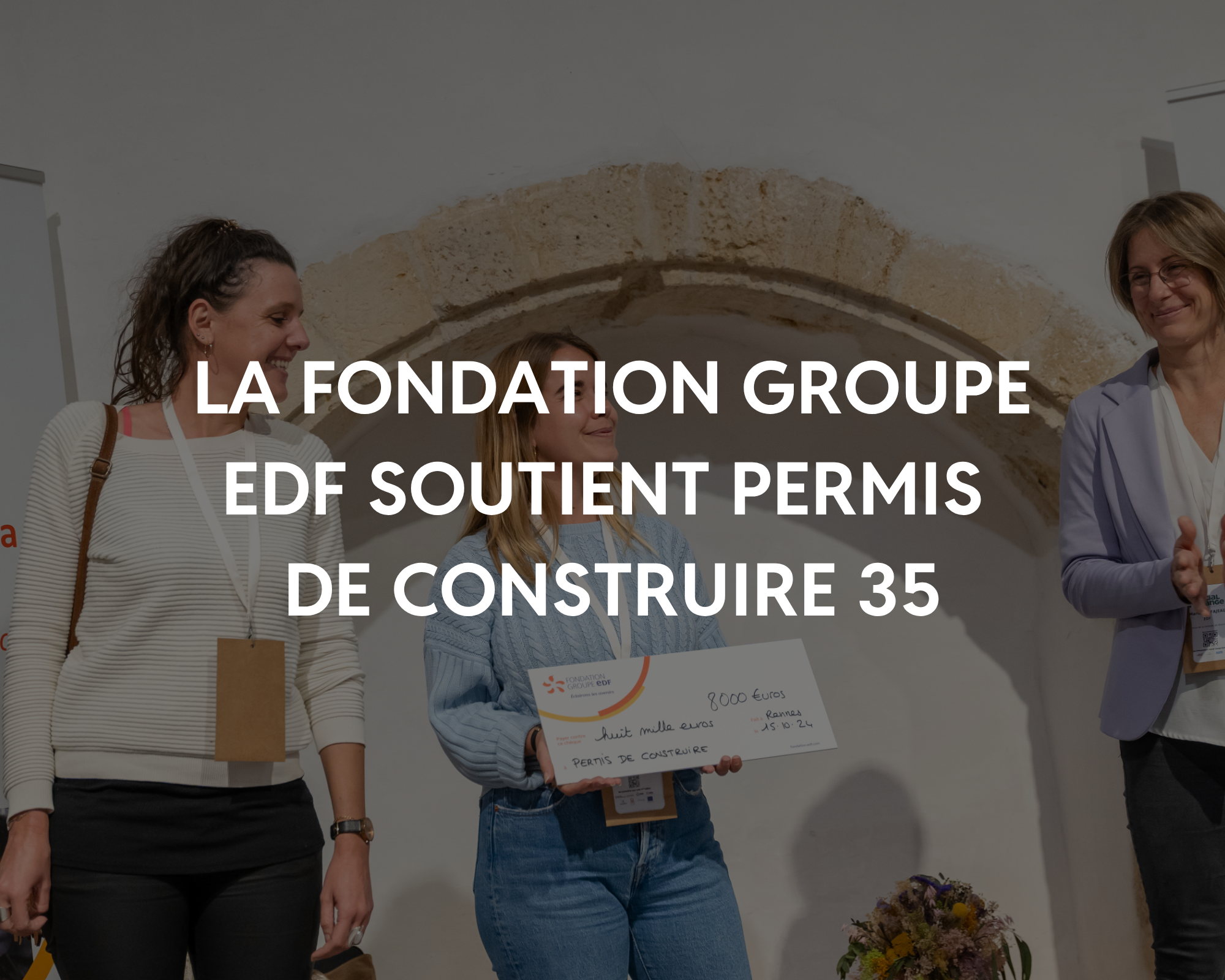 Groupe de femmes lors d'une cérémonie de soutien financier, avec un chèque à l'avant-plan, en institution avec un mur en pierre en arrière-plan.
