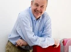 Homme souriant portant une chemise bleue, debout près d'une chaise rouge.