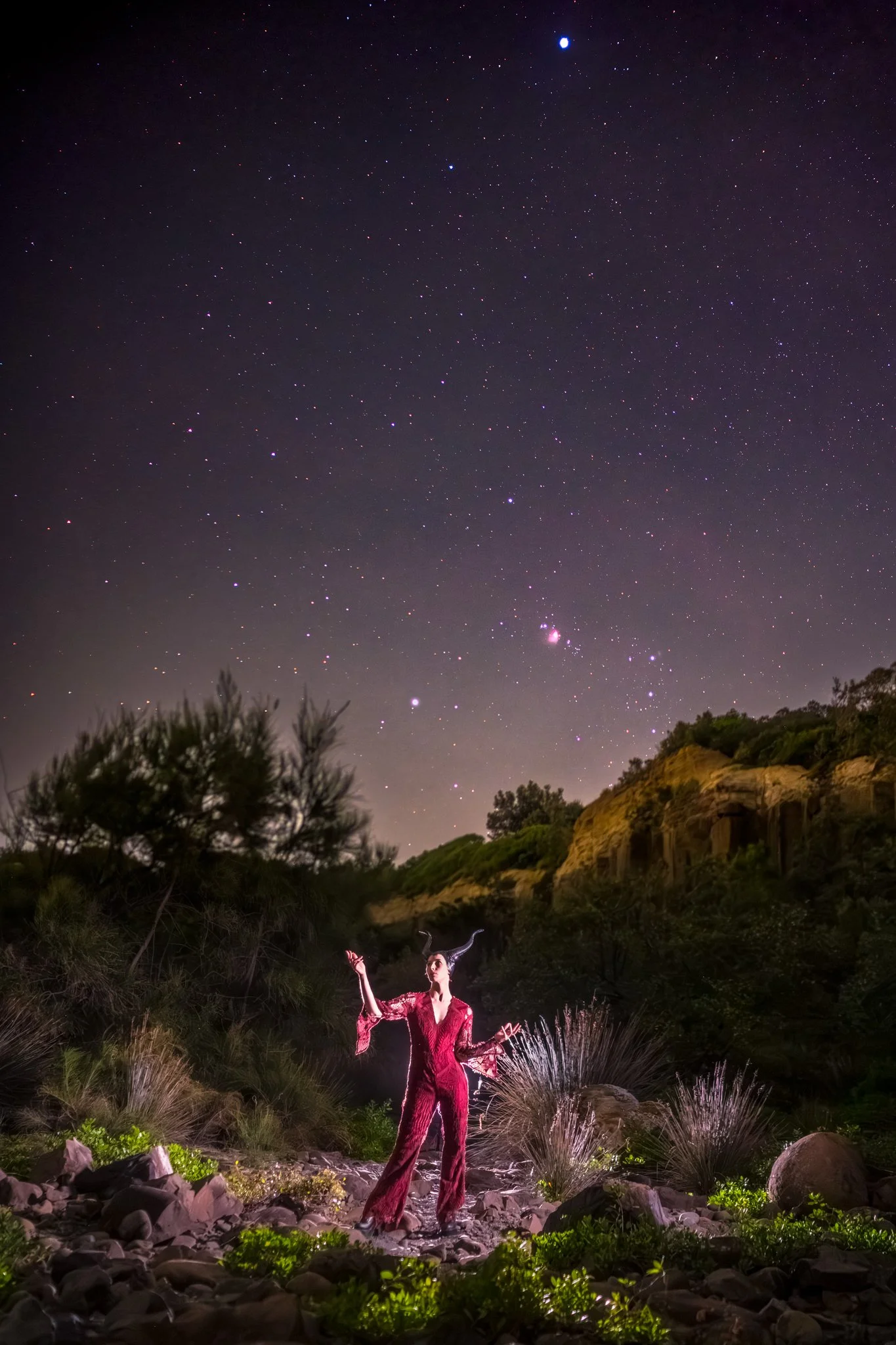 A female model standing under the night sky wearing red dress with horn with Orion as backdrop
