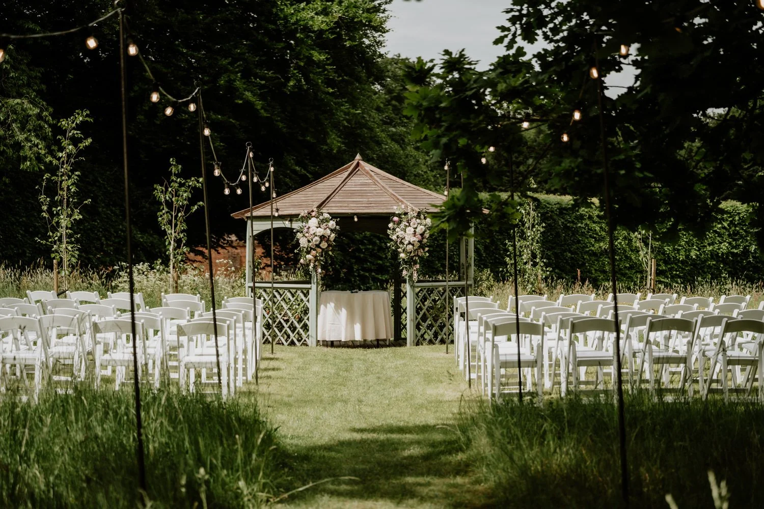 Outdoor wedding ceremony setup in orchard meadow with floral arch and white chairs at Lanwades Hall, Suffolk