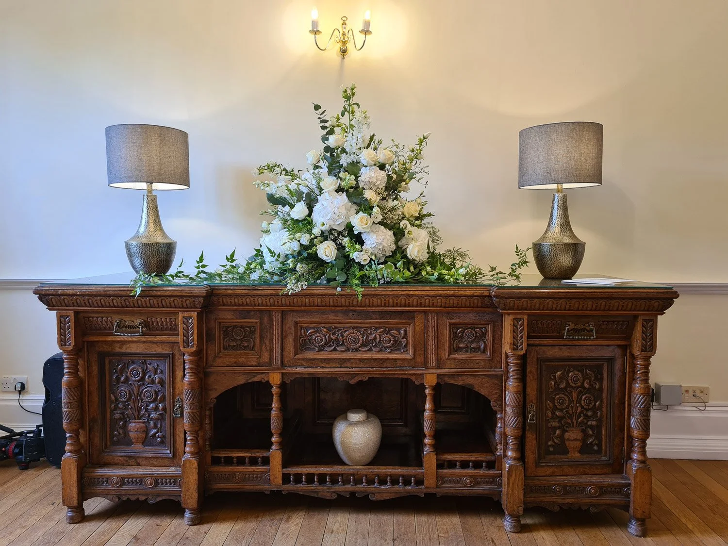 Elegant floral arrangement displayed on a carved wooden sideboard at Lanwades Hall, Suffolk, prepared for a celebration of life gathering