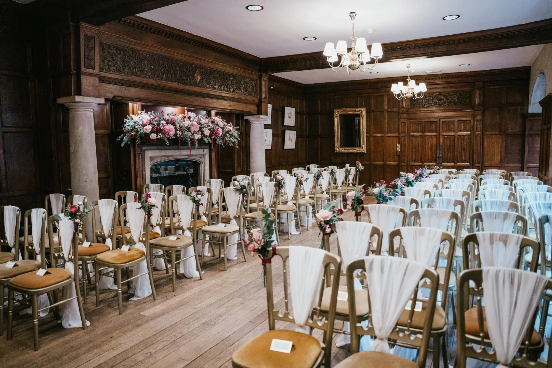 Romantic wedding ceremony setup in the wood-panelled room at Lanwades Hall, Suffolk, featuring elegant gold chairs with white drapes and a floral display of pink and red blooms above the fireplace
