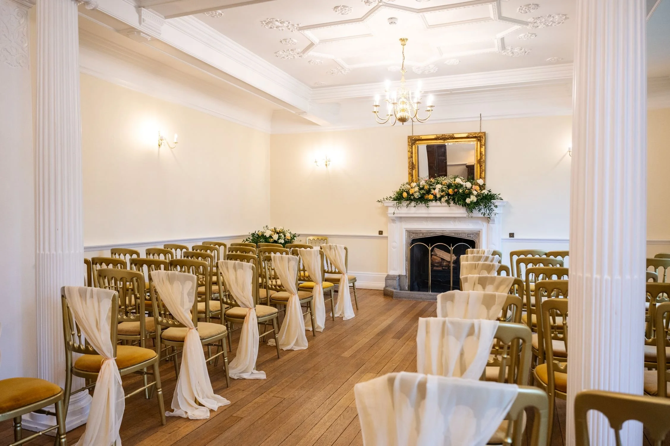 Elegant indoor wedding ceremony room at Lanwades Hall, Suffolk, featuring gold chairs with soft draping, floral arrangements, and a grand fireplace for romantic civil ceremonies