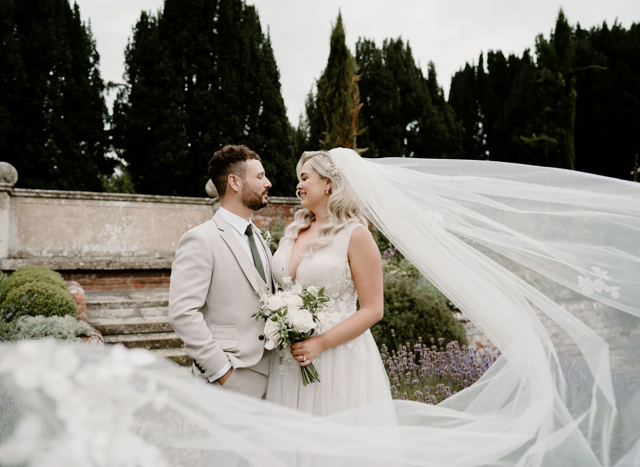 Bride and groom standing together in the gardens of Lanwades Hall, Suffolk, with the bride’s flowing veil captured in the breeze for a romantic countryside wedding portrait