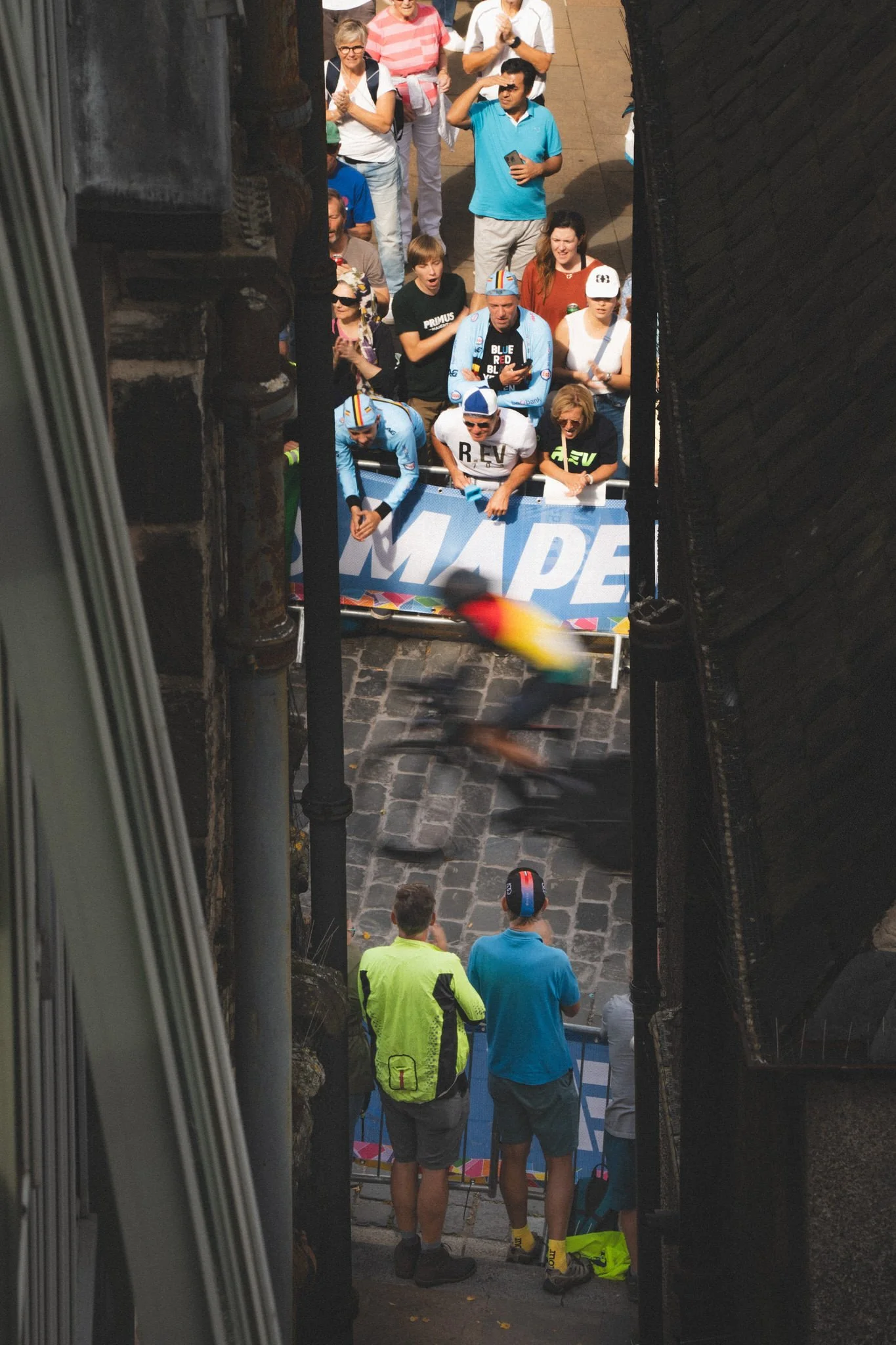 A cyclist racing through a narrow street, with spectators and officials watching from behind barriers.