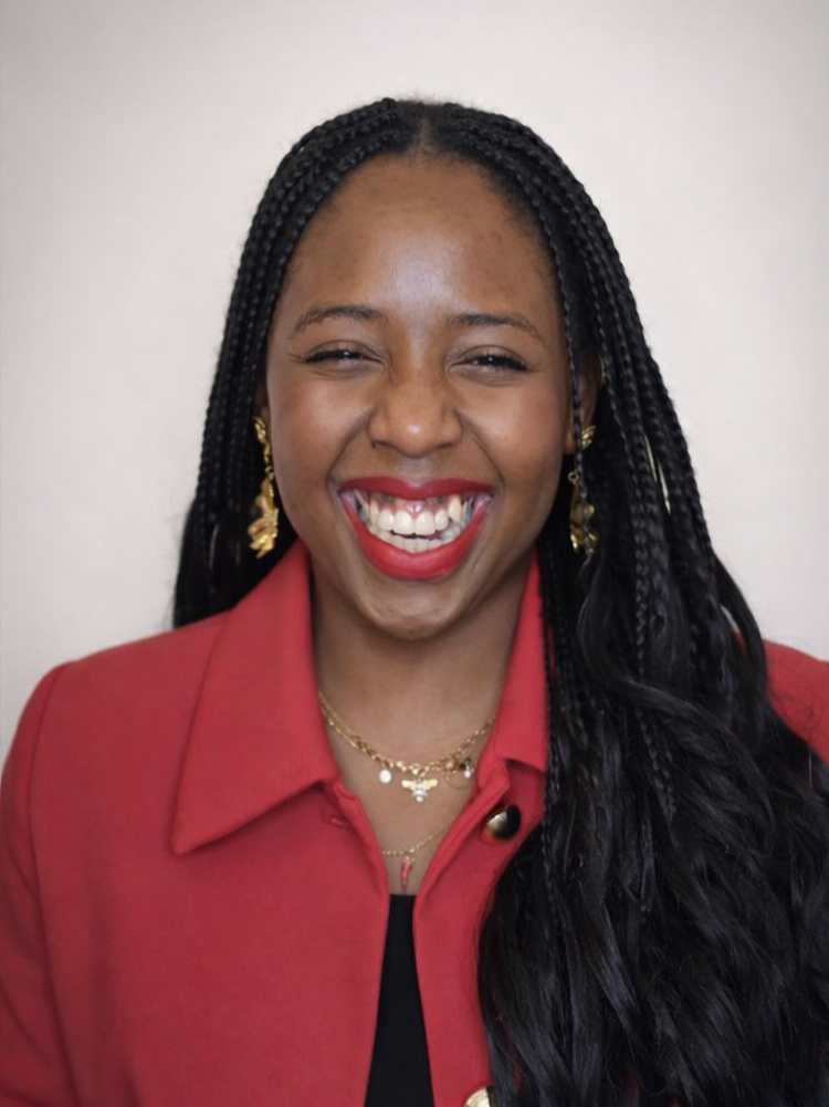 A woman smiling broadly, showing her teeth, with dark braided hair, wearing a red blazer, gold jewelry, and red lipstick against a plain background.