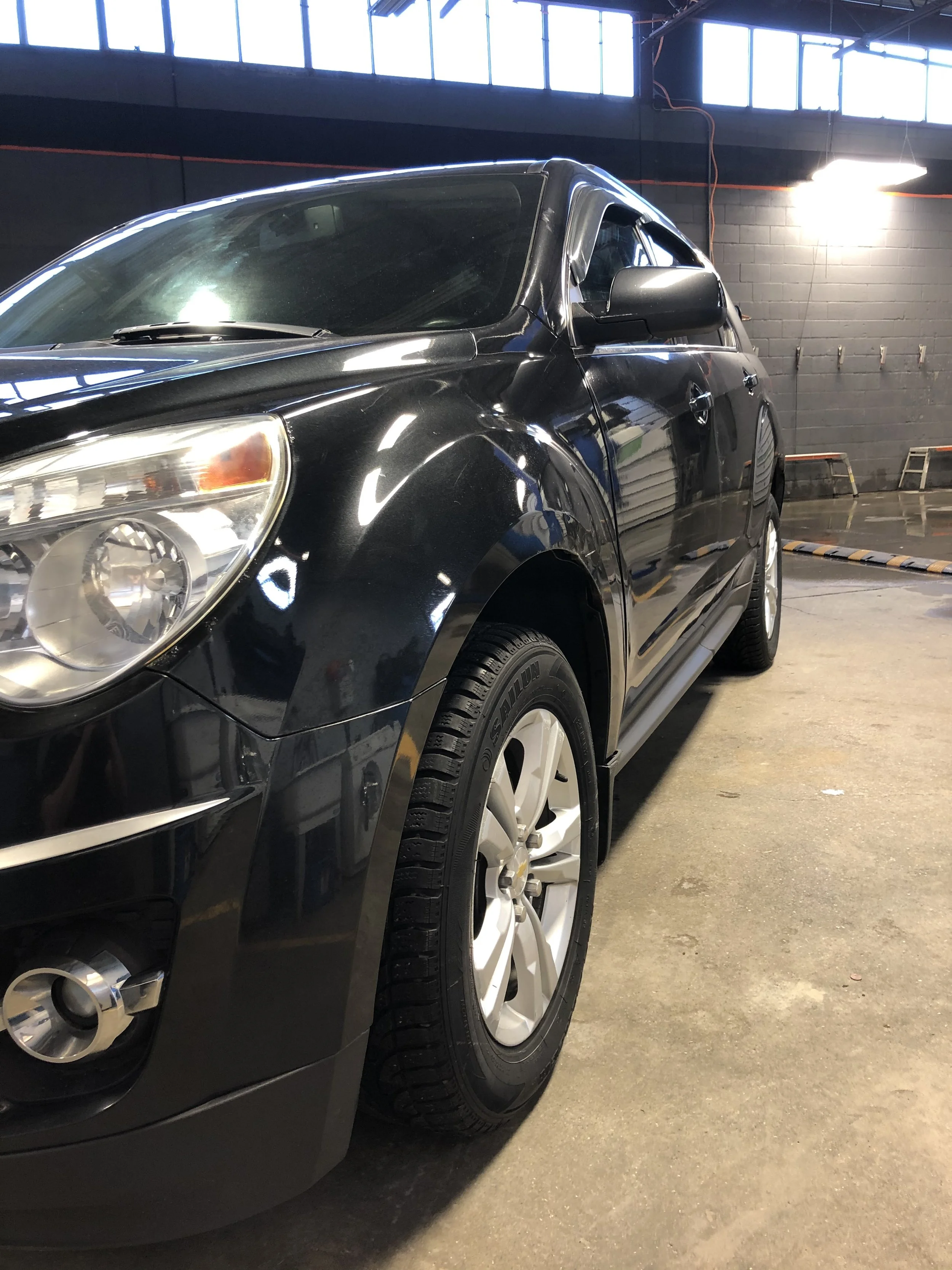 Black SUV parked inside a garage with concrete floor and dark brick walls, overhead fluorescent lights, and large windows at the top.