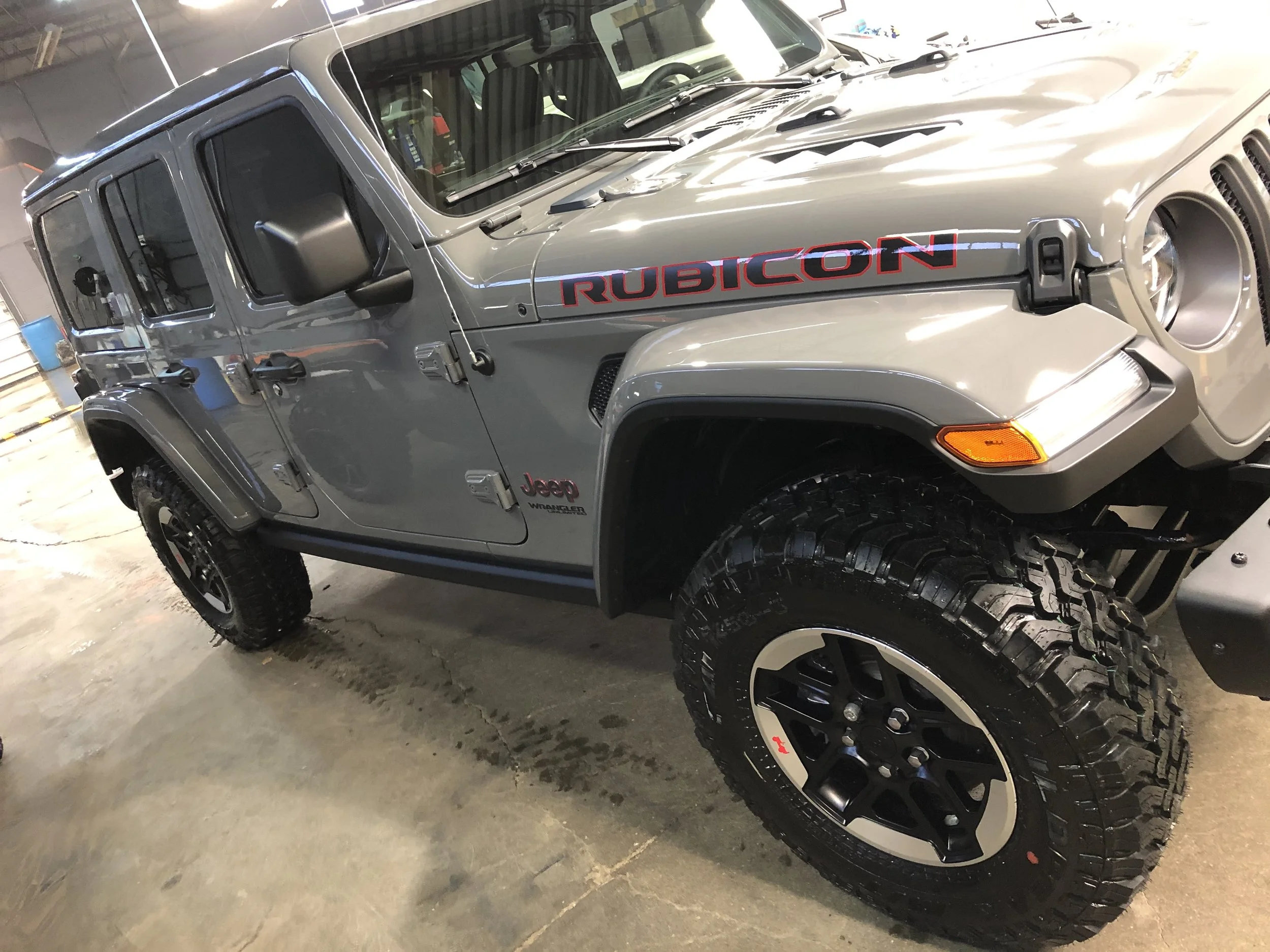 A silver Jeep Rubicon parked indoors with large off-road tires and black rims, featuring a black side mirror, and red and black decals on the hood.