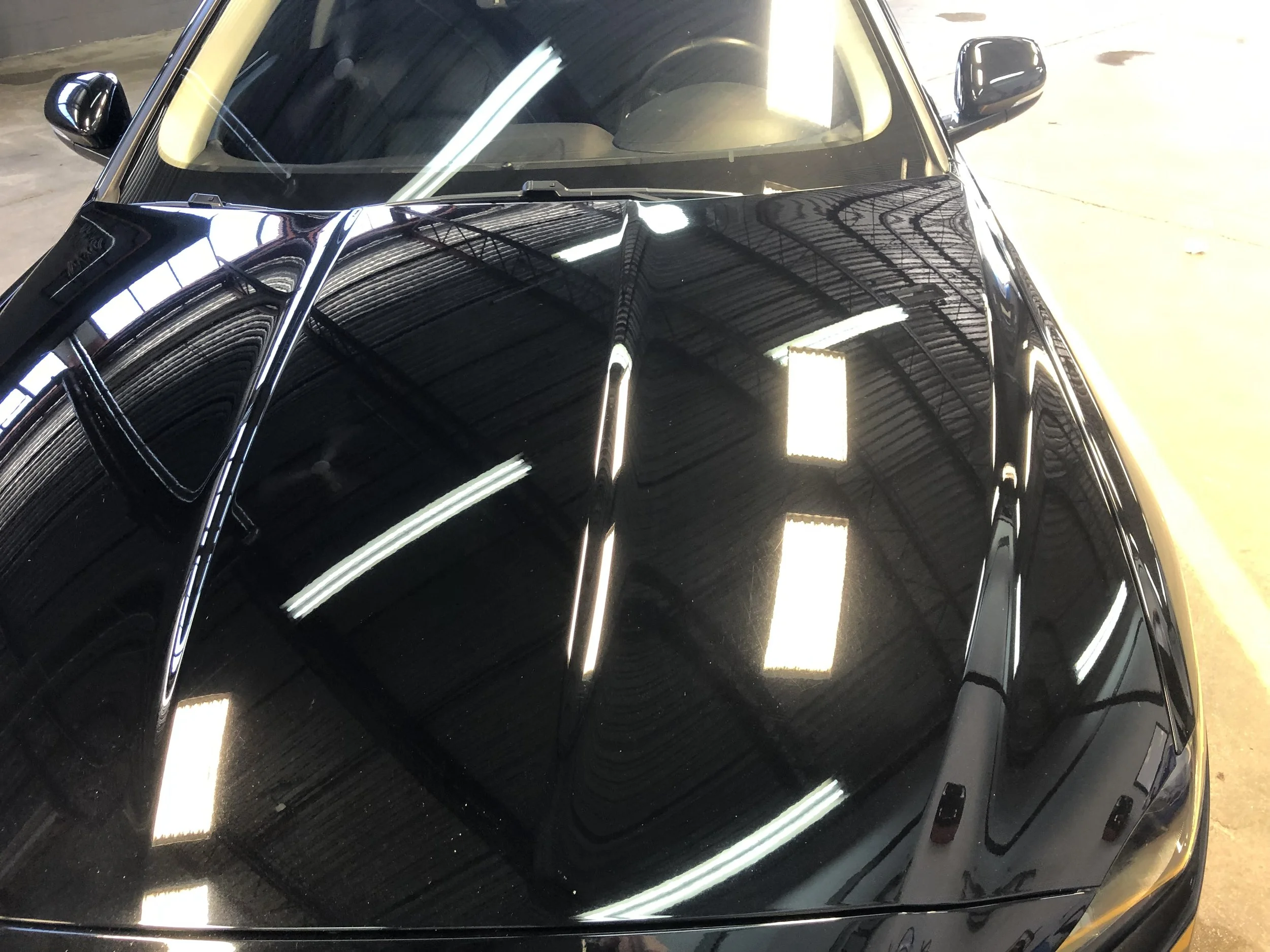 Close-up of a black car's hood with reflective surface showing overhead fluorescent lights and a warehouse ceiling.