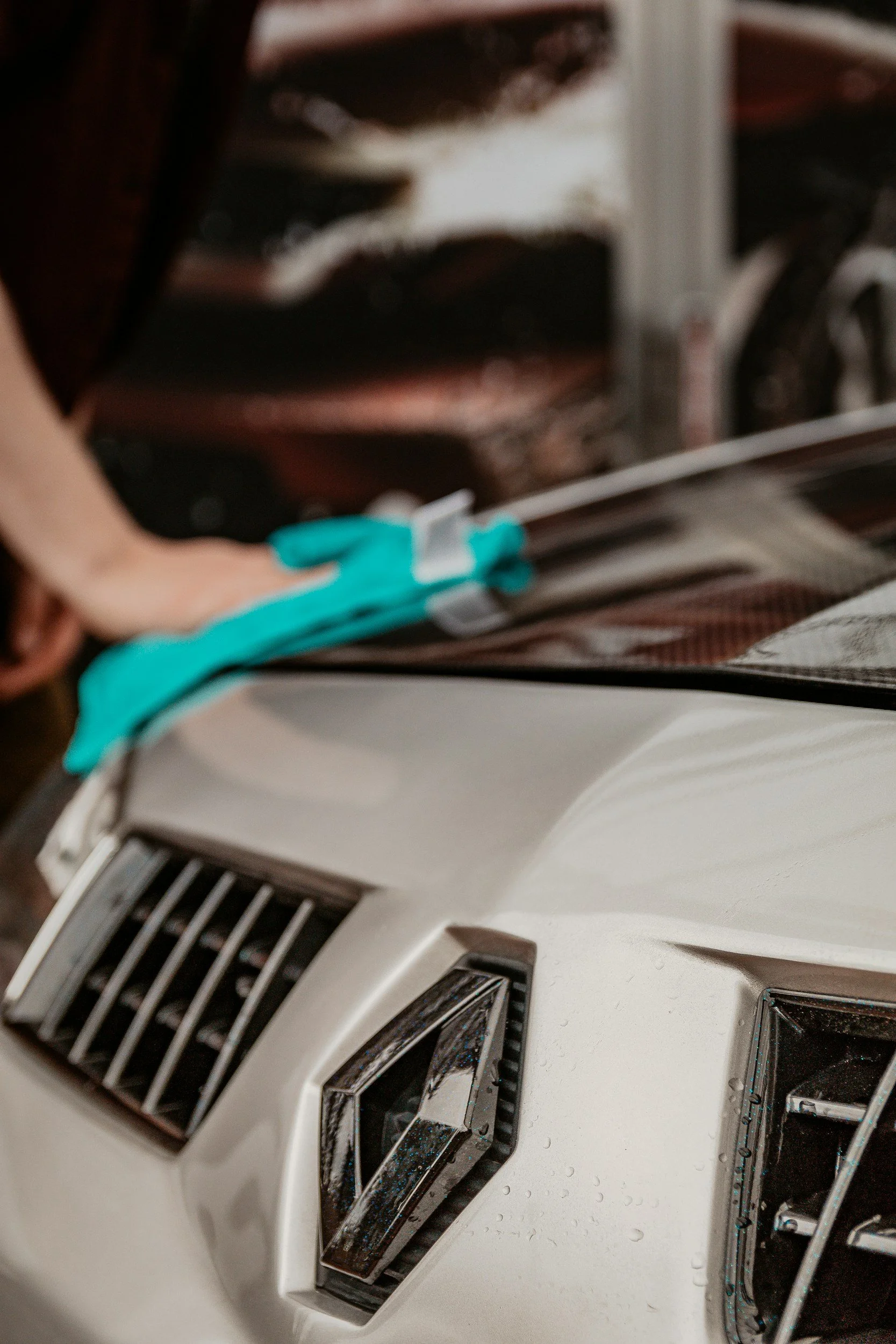 Close-up of a white car's front section, showing the Renault logo on the grille, with water droplets on the surface, and a person's hand in the background holding a cloth.
