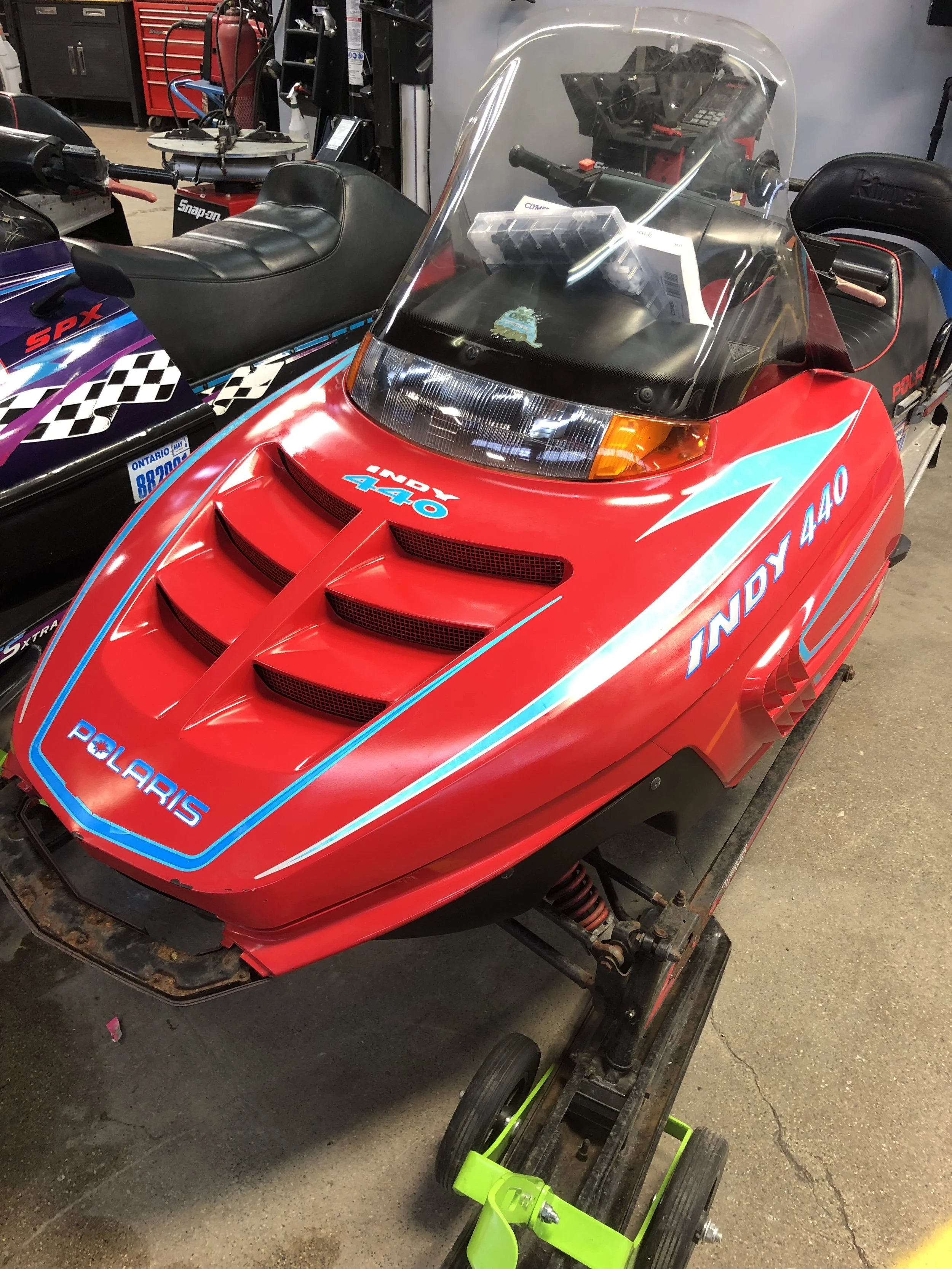 A vintage red Polaris Indy 440 snowmobile with blue and white decals, parked indoors on a black dolly, next to another snowmobile and workshop tools.