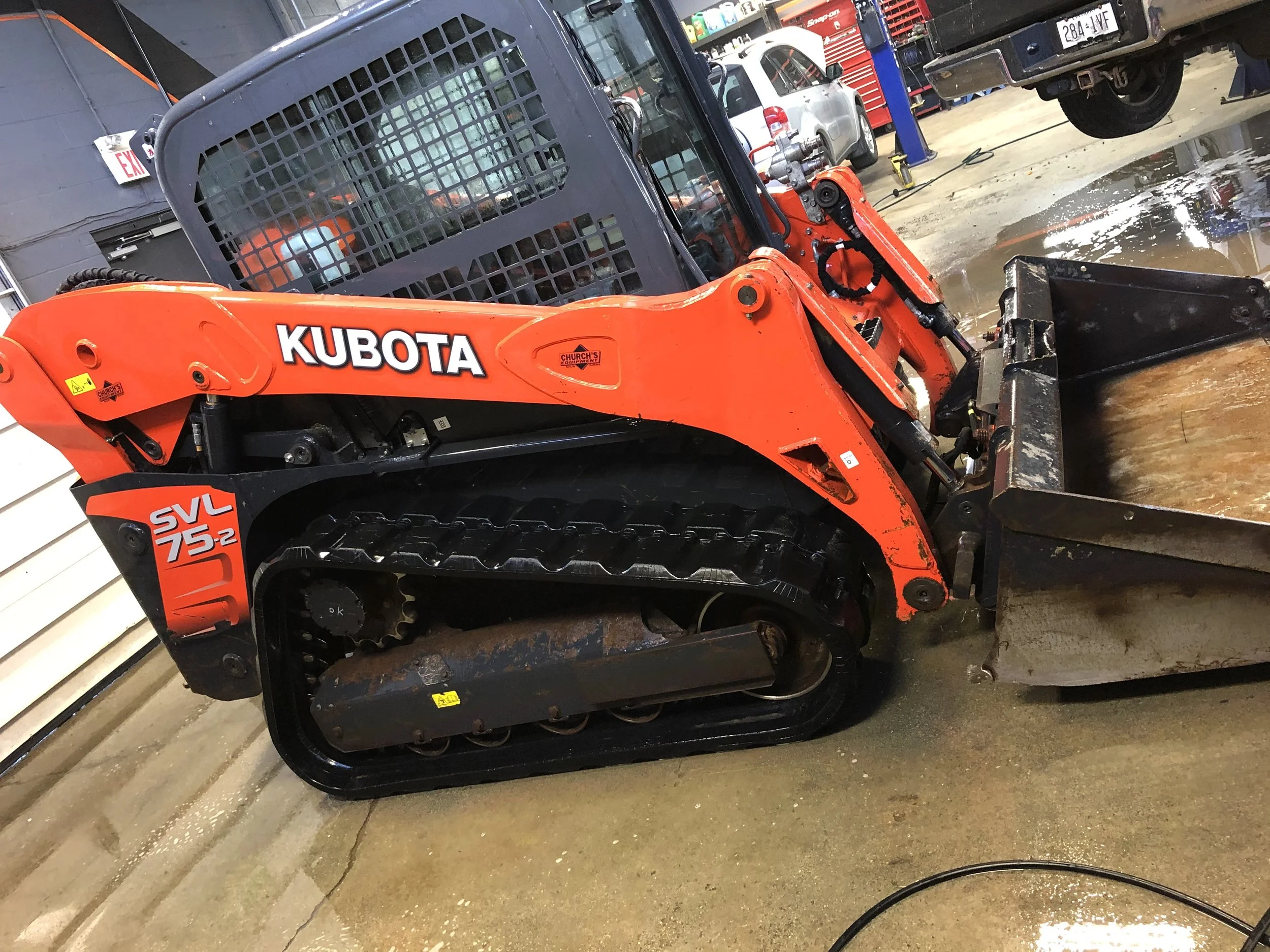 A Kubota SVL75-2 compact track loader in orange and black, parked indoors with a wet floor, with various vehicles and equipment in the background.