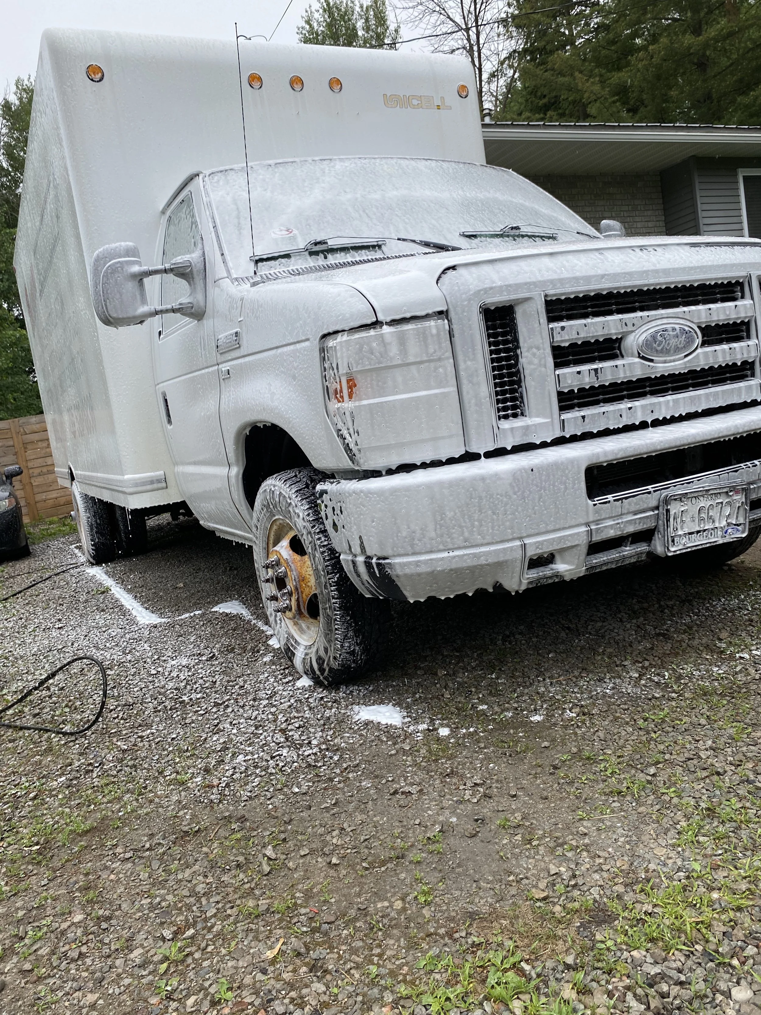 White delivery truck covered in soap suds during a wash outside on a gravel driveway.