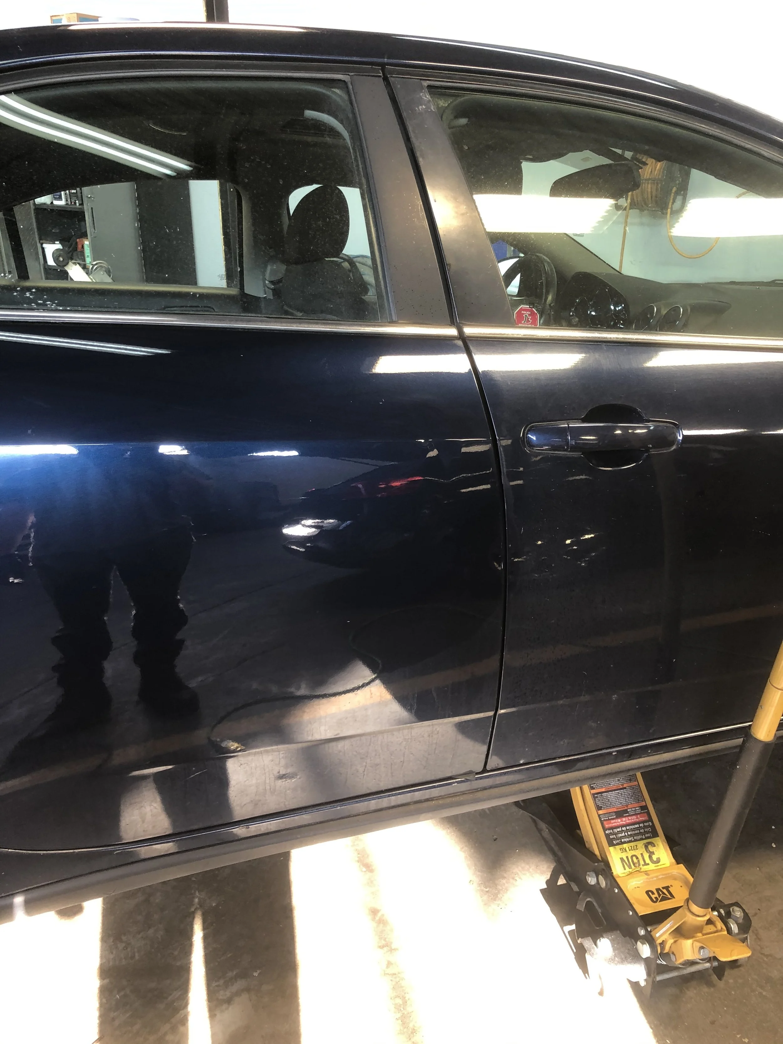 Close-up of a black car inside a garage, showing the driver's side door and window, with a yellow hydraulic jack underneath the car.