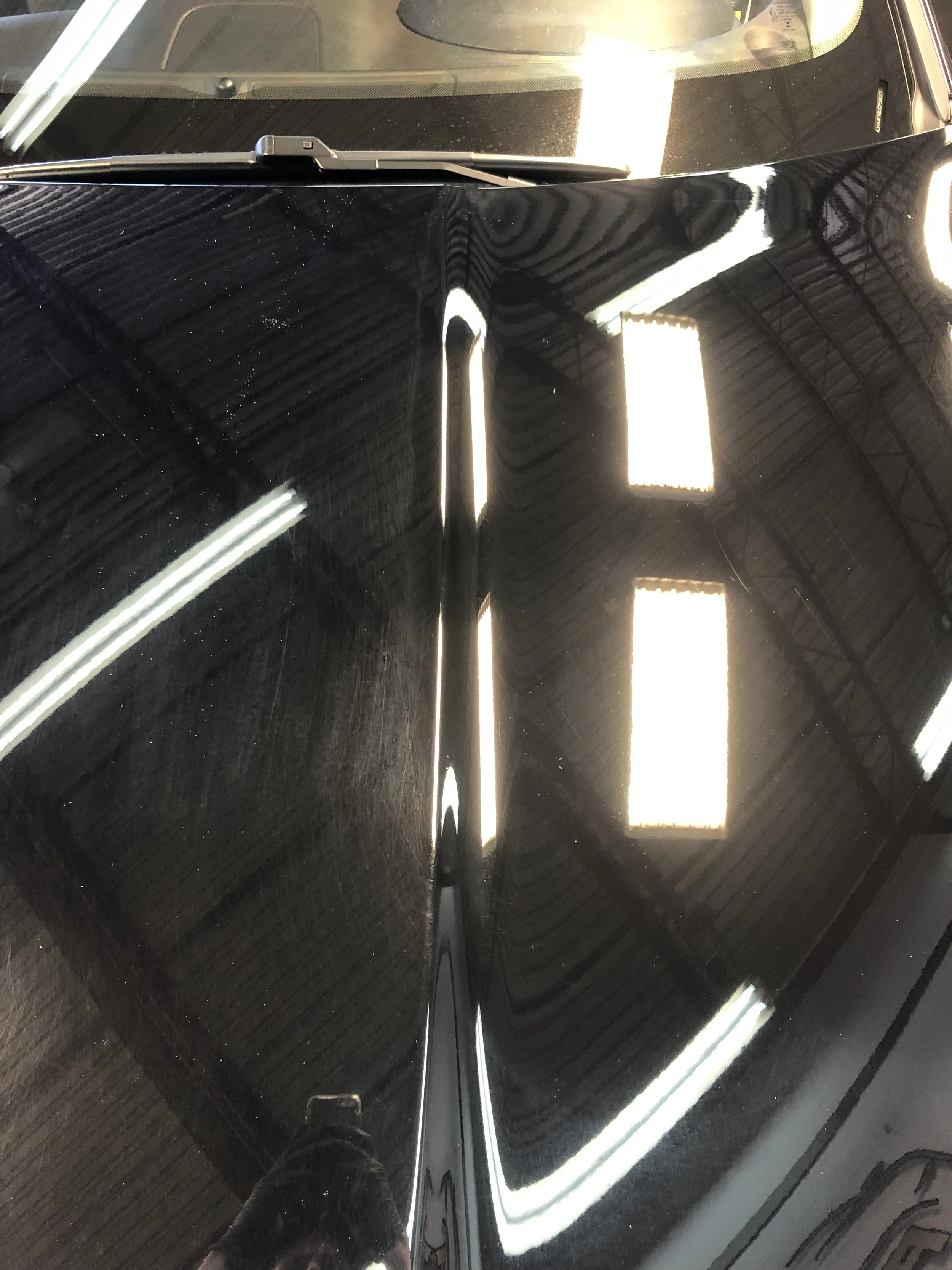 Close-up of a black car's hood reflecting overhead ceiling lights and part of the surrounding environment.