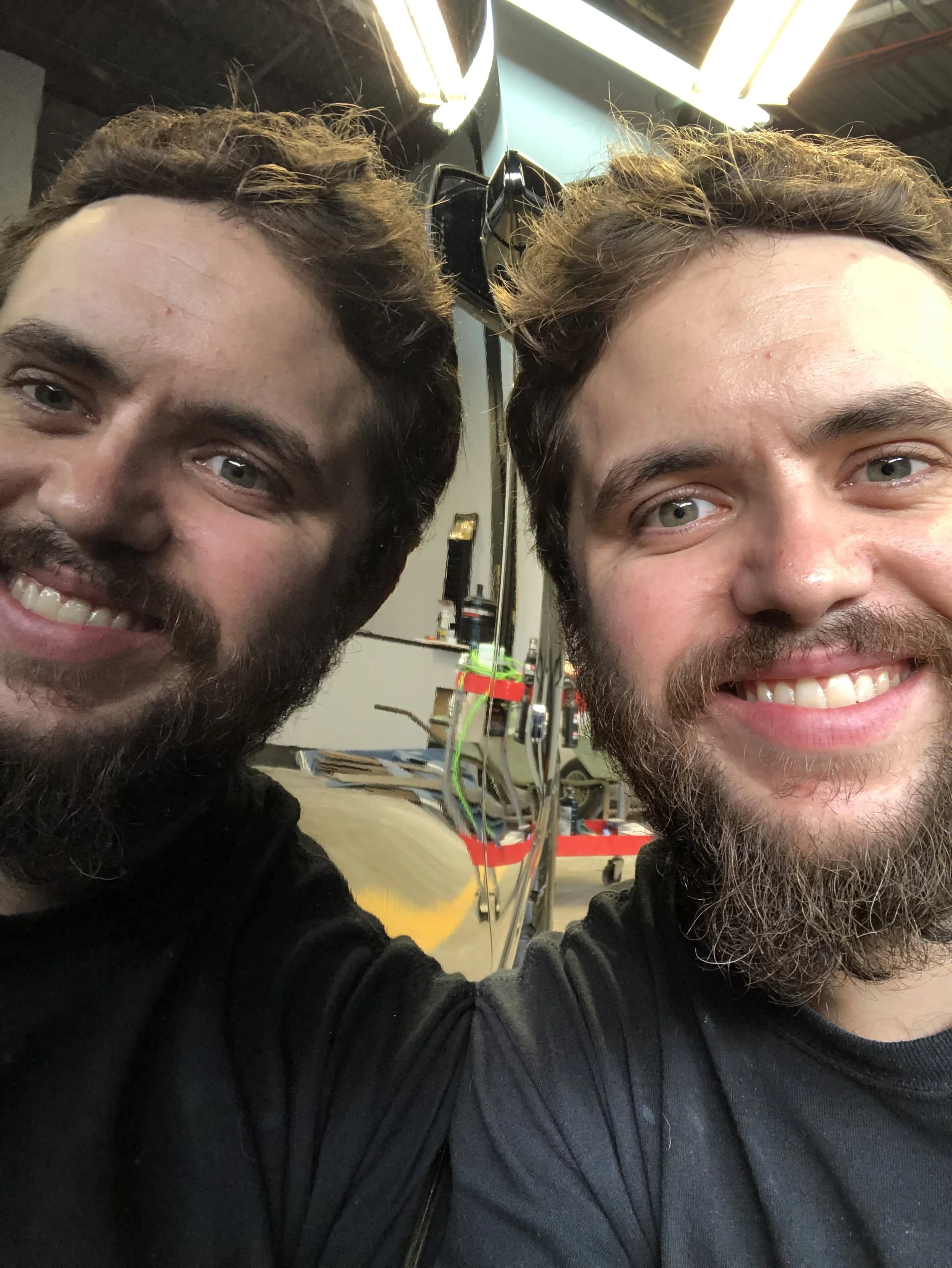 Two men with beards smiling and taking a selfie inside a workshop or studio with equipment and tools in the background.