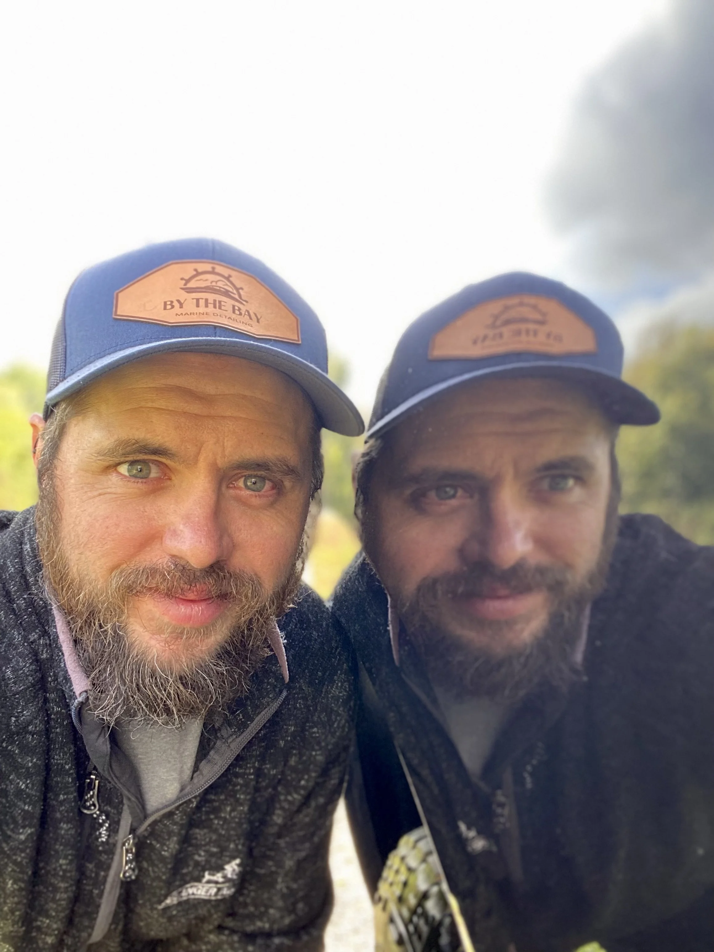 Two men with beards wearing matching blue caps and black jackets taking a close-up selfie outdoors on a cloudy day with trees in the background.