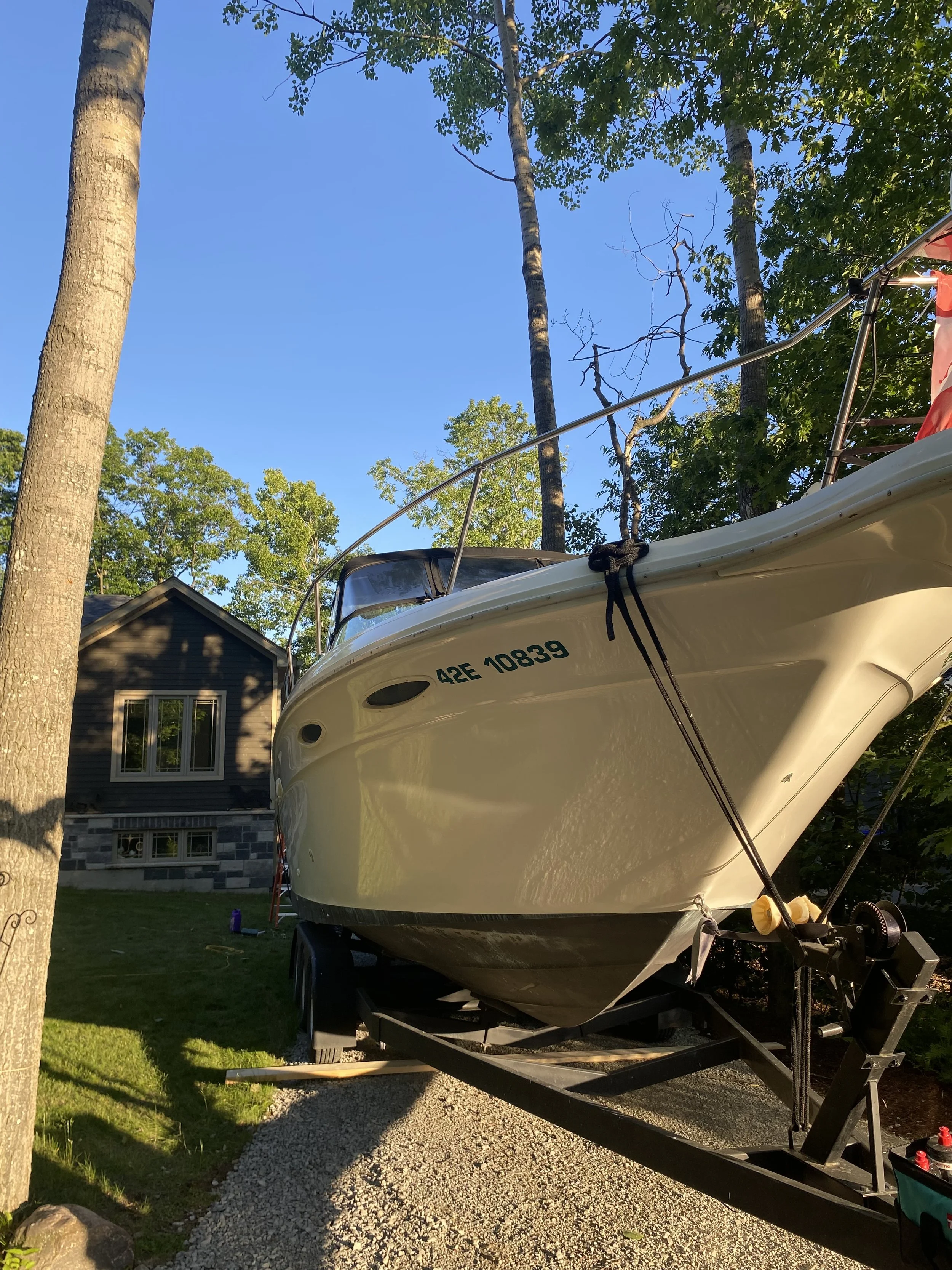 A white boat on a trailer parked on gravel in a backyard with trees and a house in the background.
