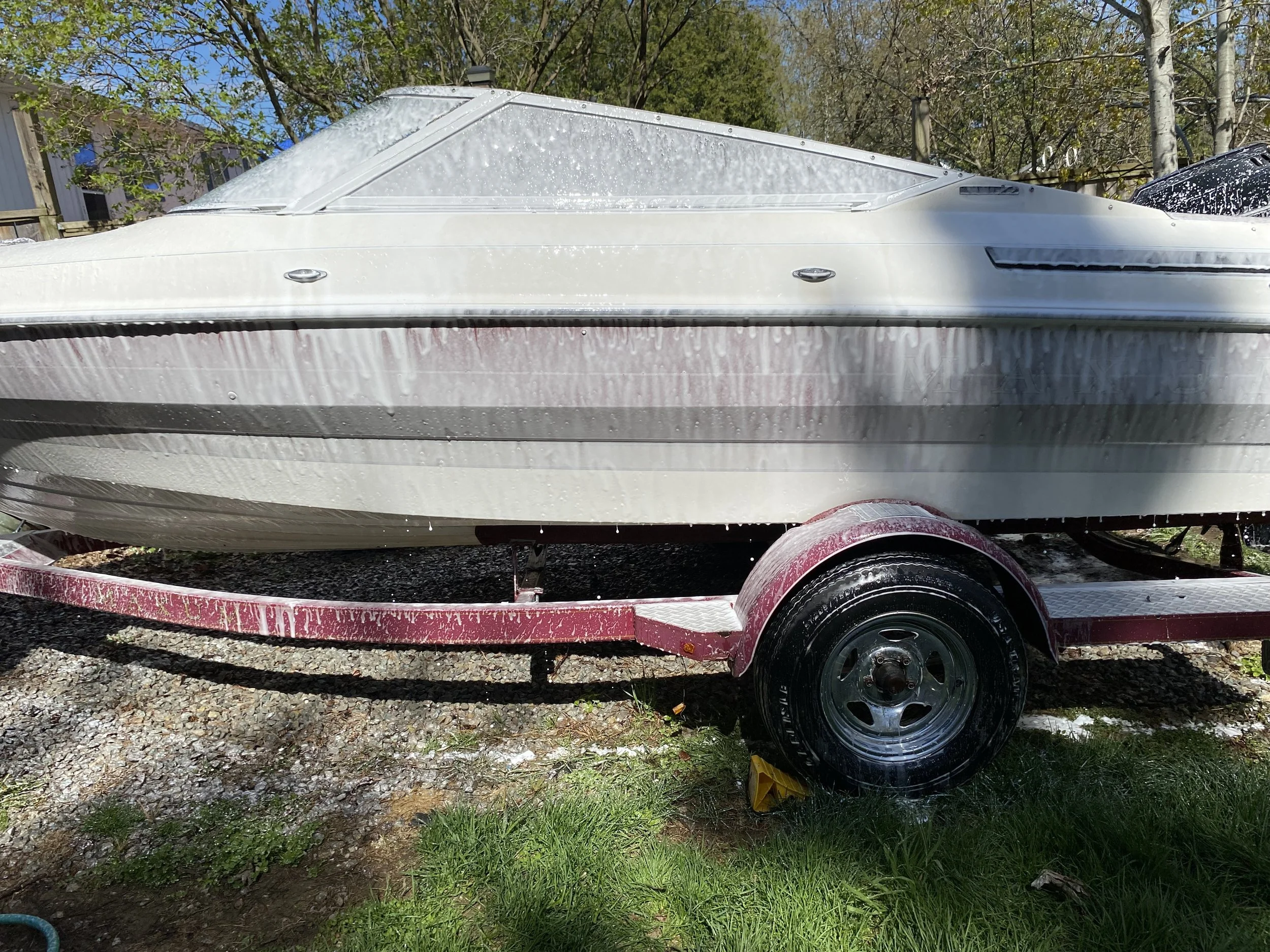 A boat on a pink trailer with water droplets on it, parked on grass and gravel, with trees and buildings in the background.