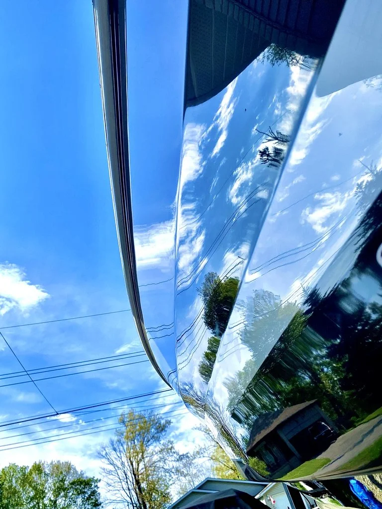 Close-up of a shiny, reflective black car hood with reflections of blue sky, clouds, trees, power lines, and a house.