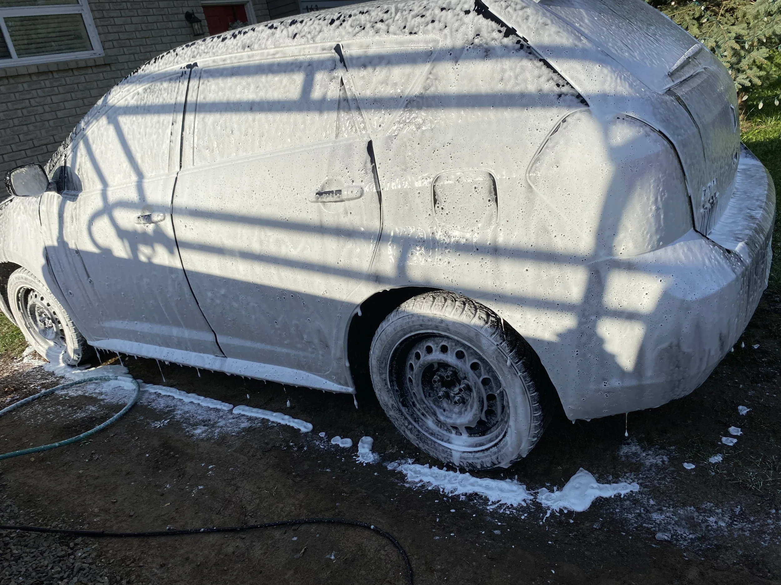 A small white car covered in soap suds during a wash, with foam dripping from its sides and wheel wells, on a dirt driveway beside a house with a brick exterior.