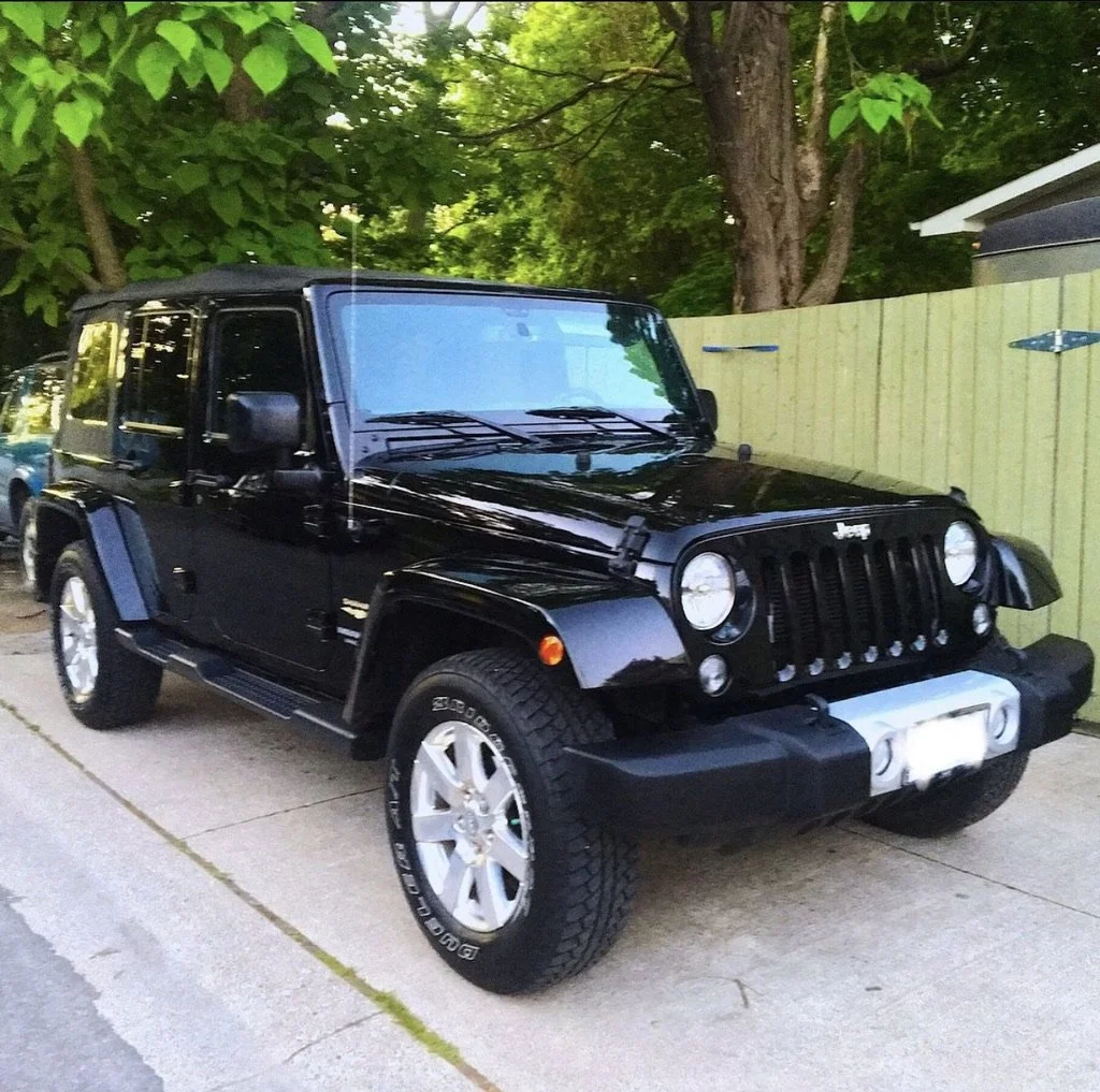 A black Jeep Wrangler parked on a driveway with green trees and a wooden fence in the background.