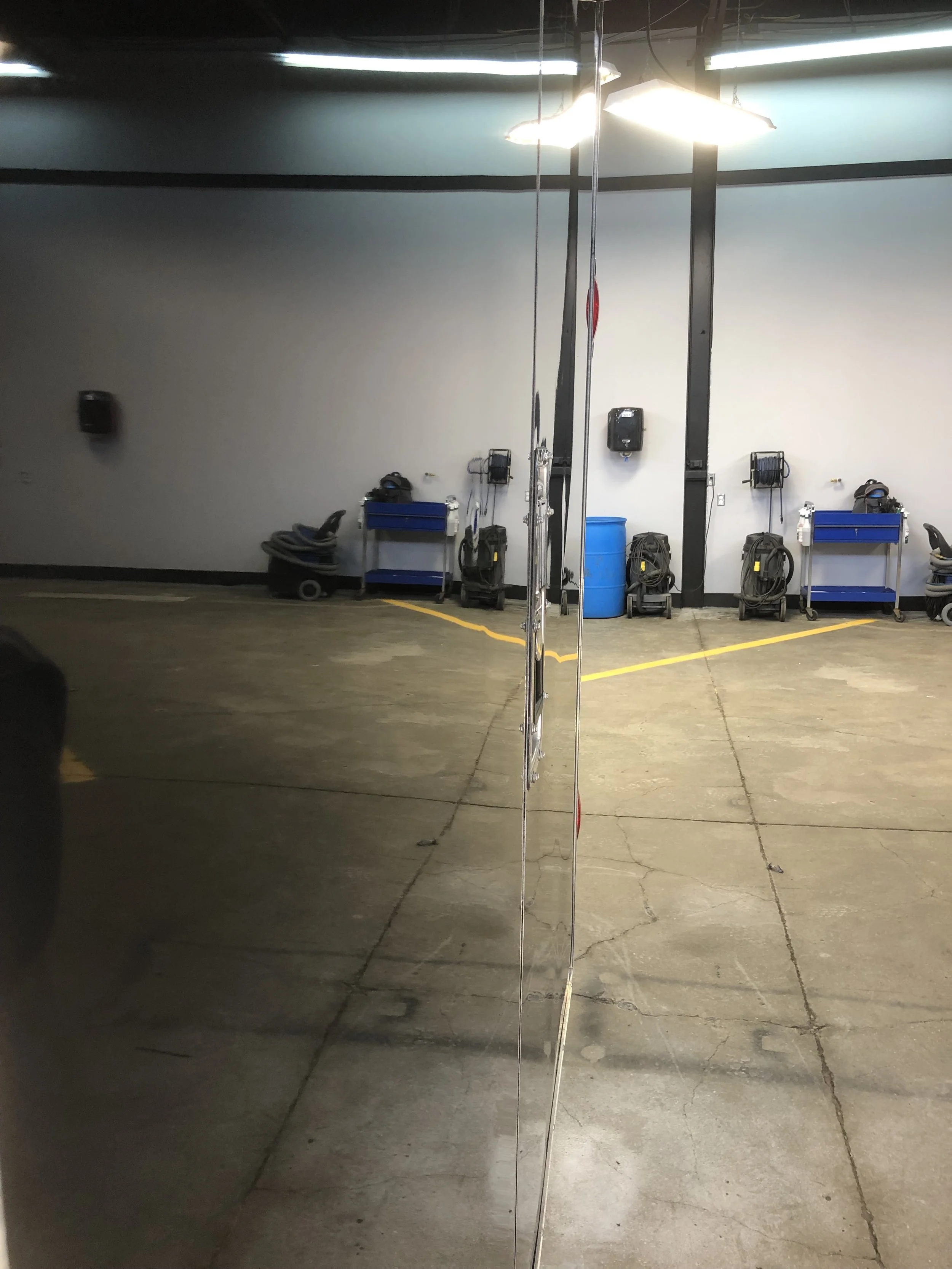 Reflection of a garage or maintenance area with equipment and tools against the white wall, including vacuum cleaners, blue carts, and a large blue barrel, with overhead fluorescent lights.