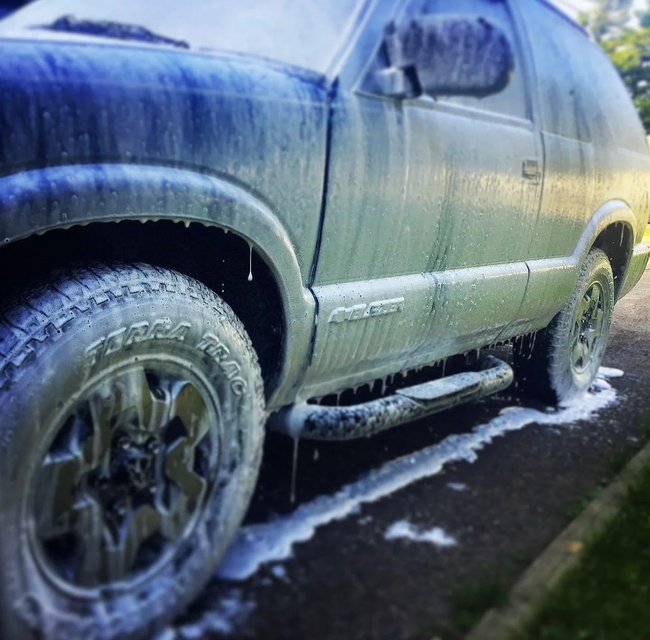 A dark-colored SUV being washed with soap and water, with soap suds and foam on the tires, sides, and ground.