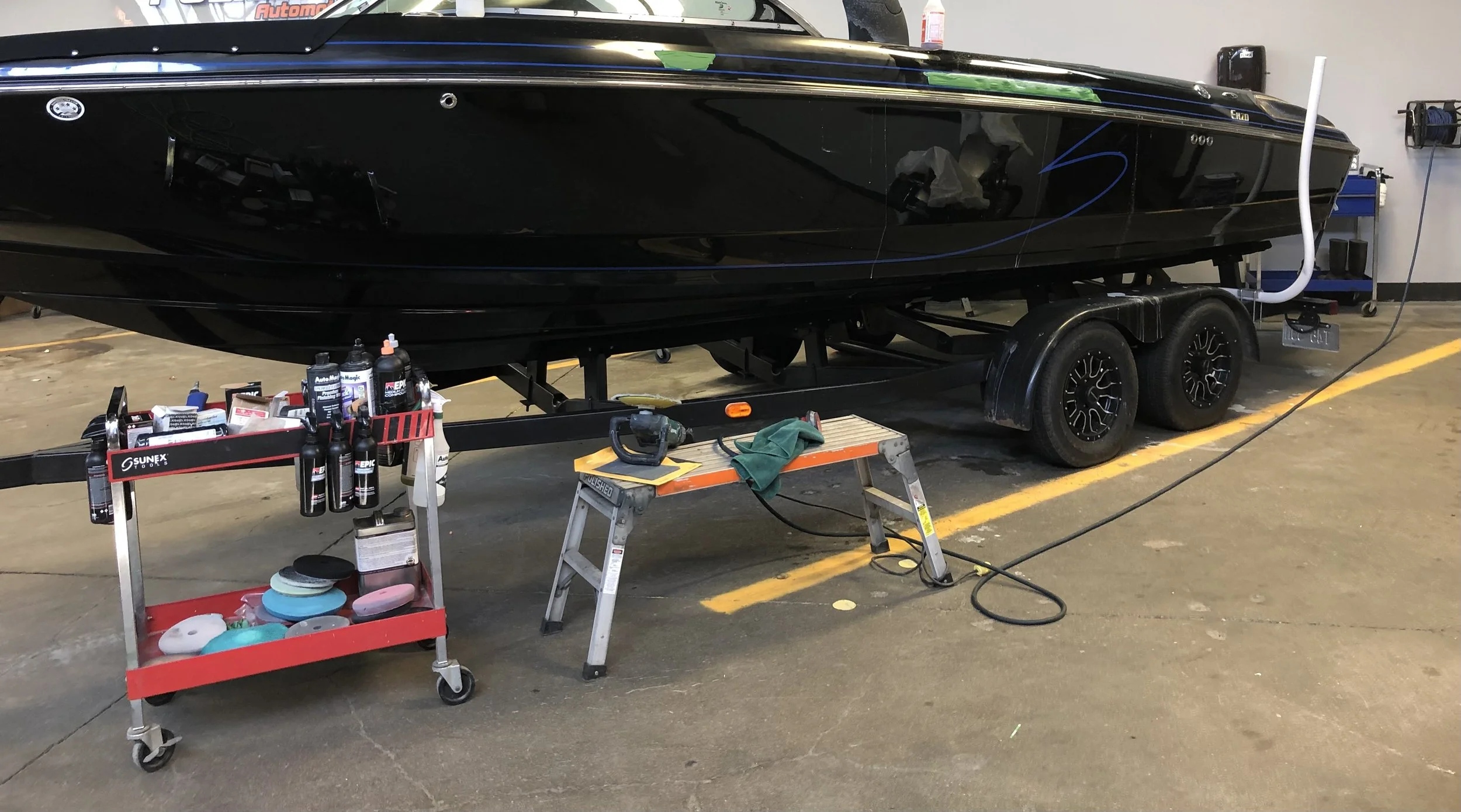 A black boat on a dual-axle trailer inside a garage with cleaning supplies, a saw, and towels nearby.