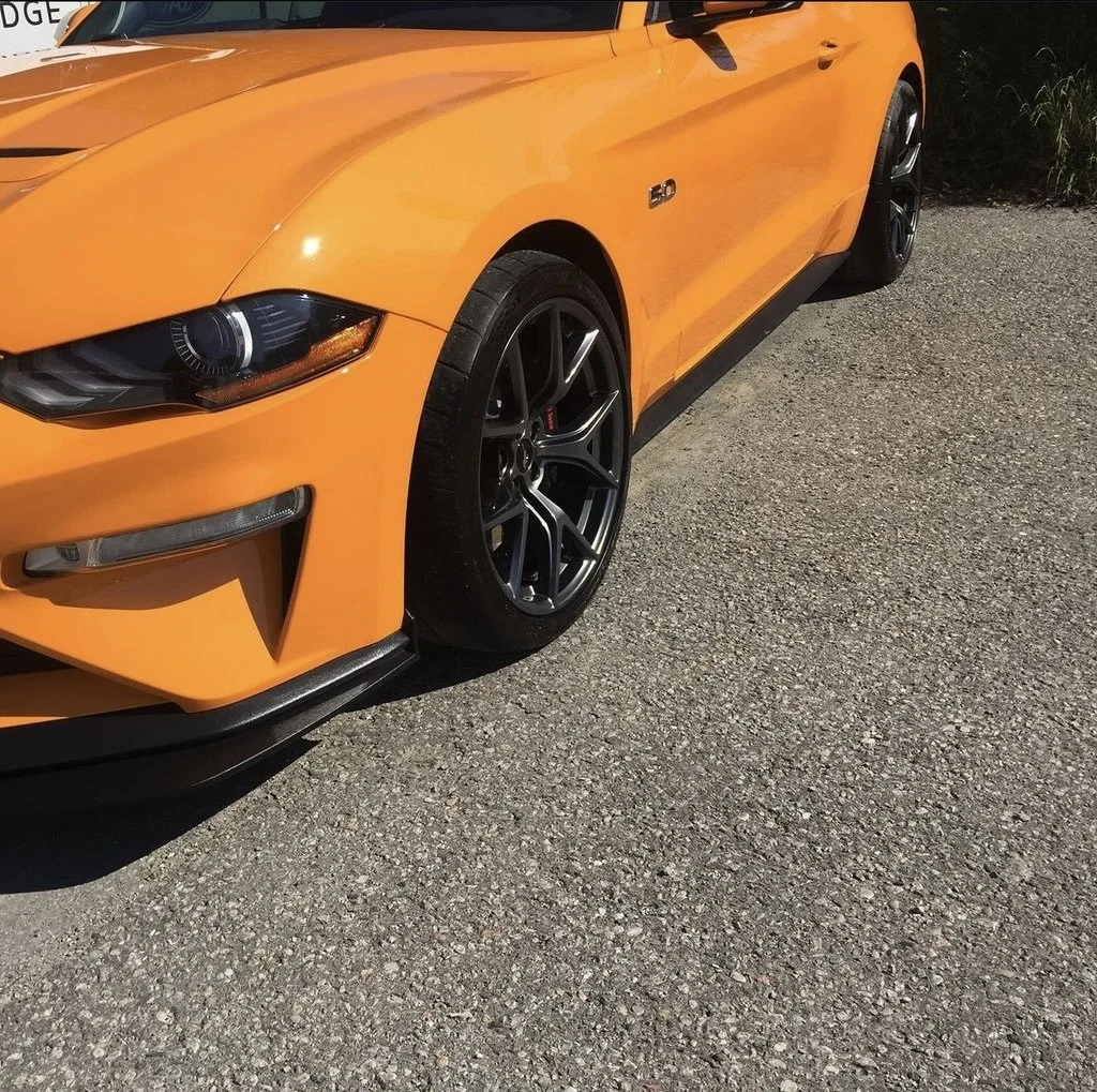 Close-up of an orange sports car parked on a gravel surface, showing the front left side including the headlight, wheel, and part of the hood.
