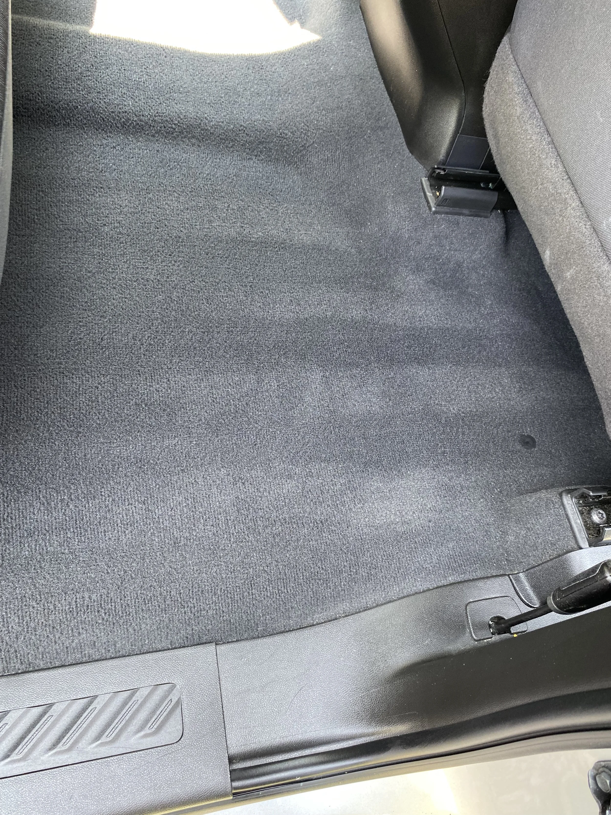 Rear passenger side floor of a car with dark carpet, part of the seat and the car door hinge visible.